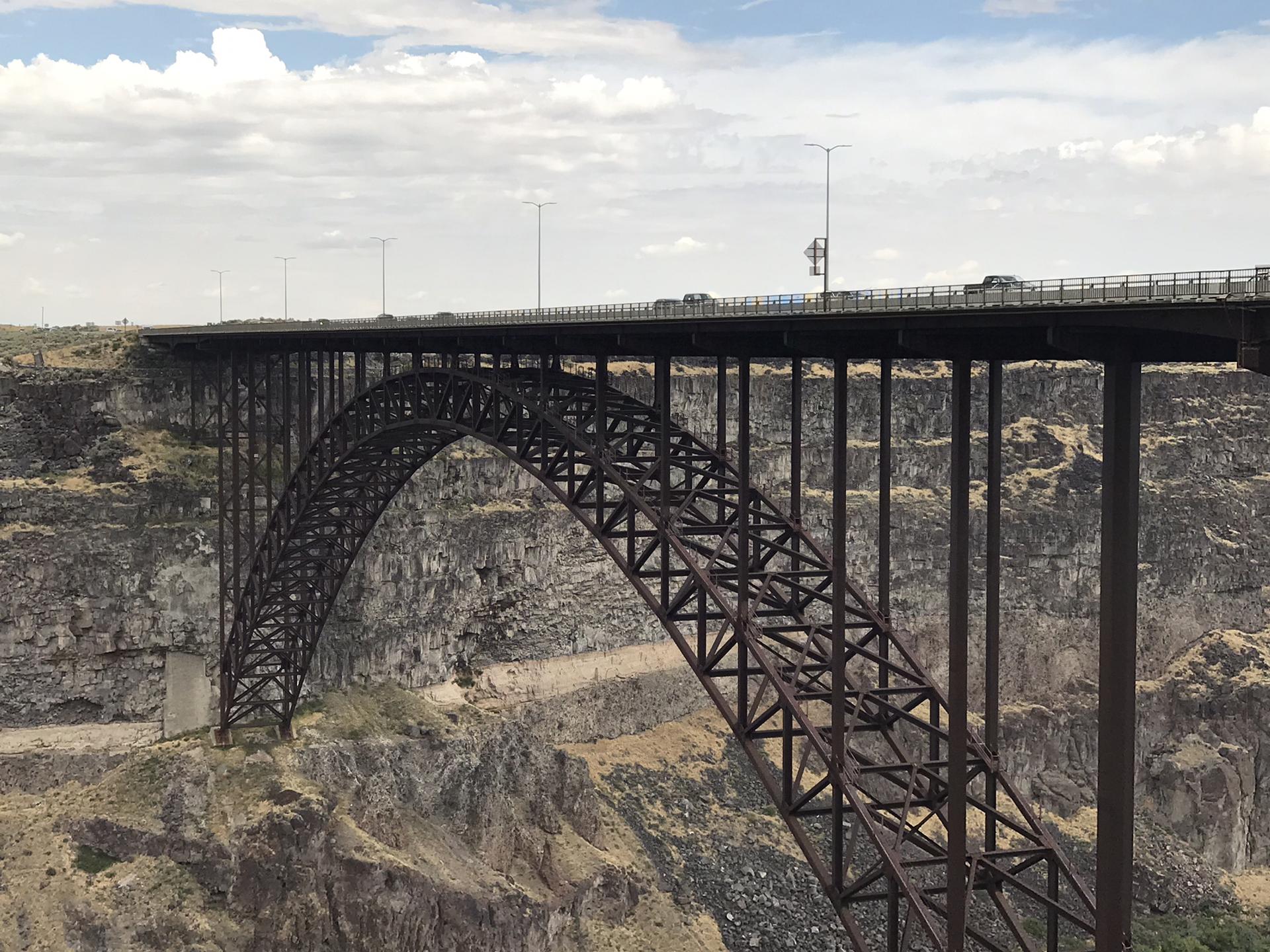 A massive steel arch bridge spans a deep, rocky canyon on a cloudy day.
