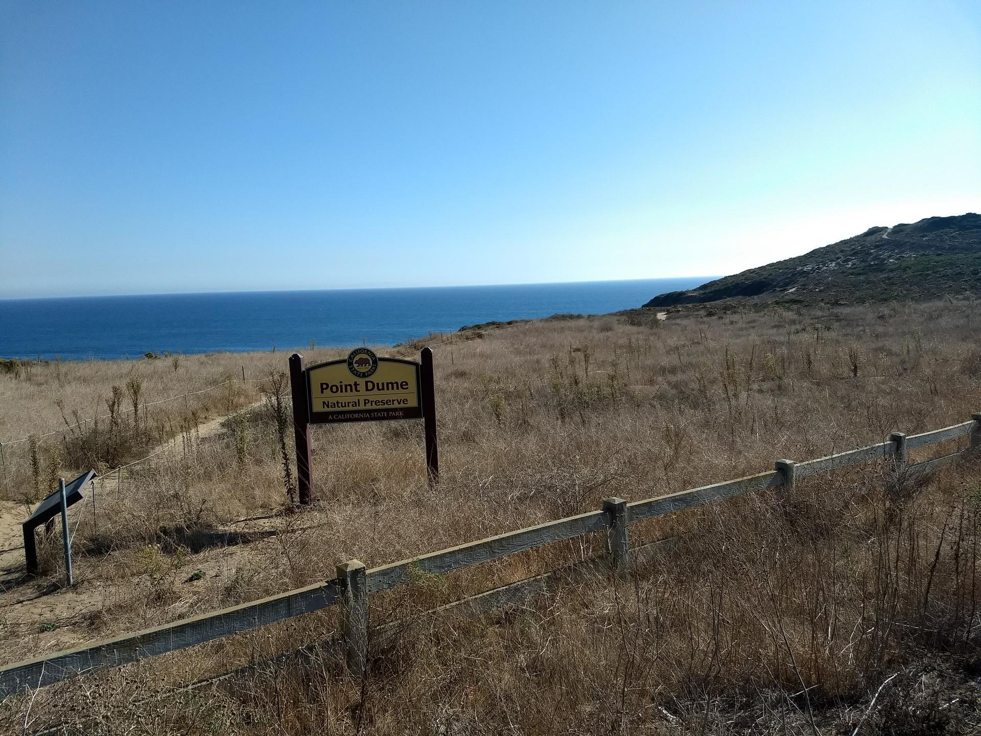 Wooden sign in dry grassy field, ocean in background, under a clear blue sky.
