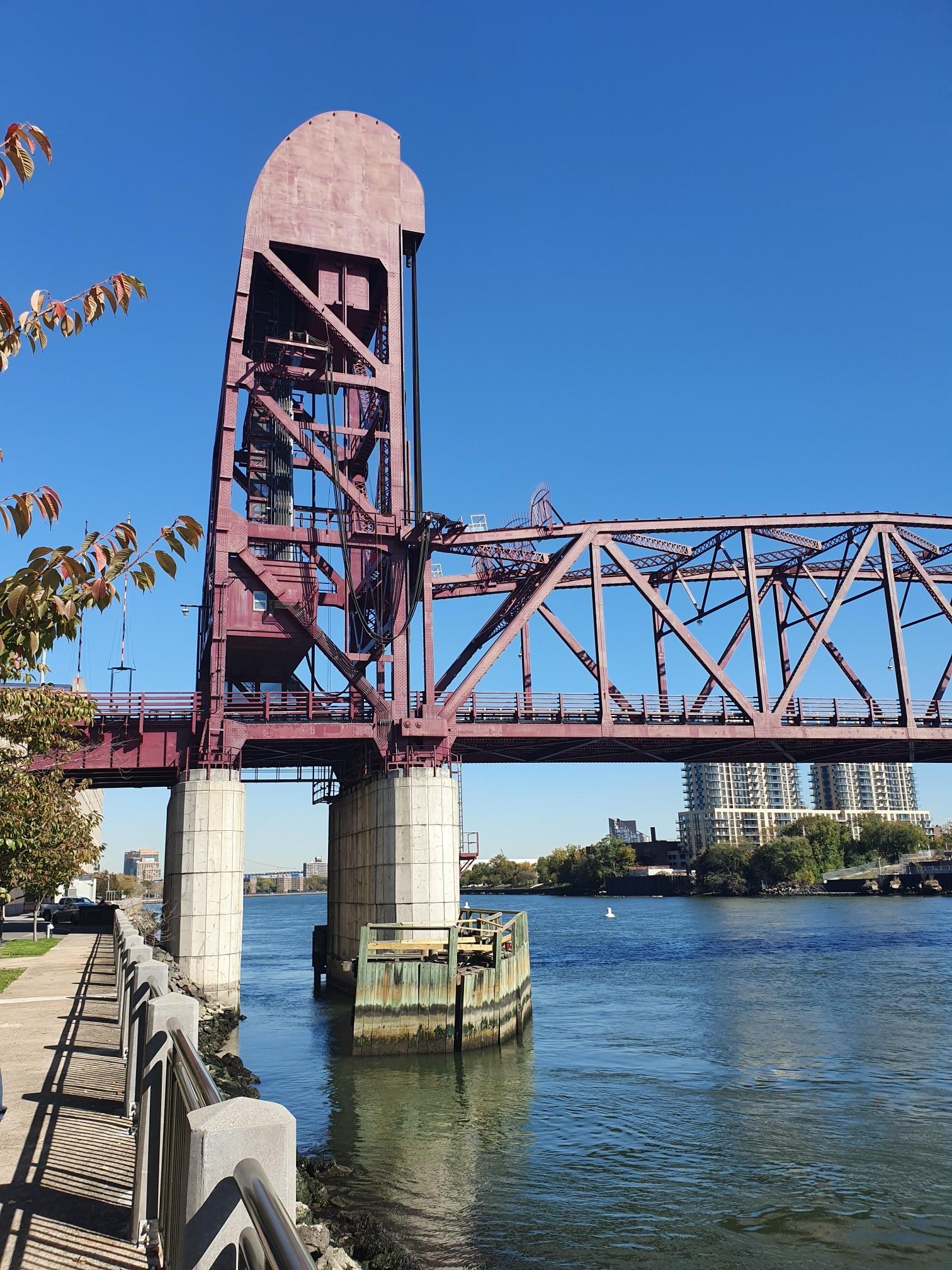 A large red metal bridge spans a river under a clear blue sky.