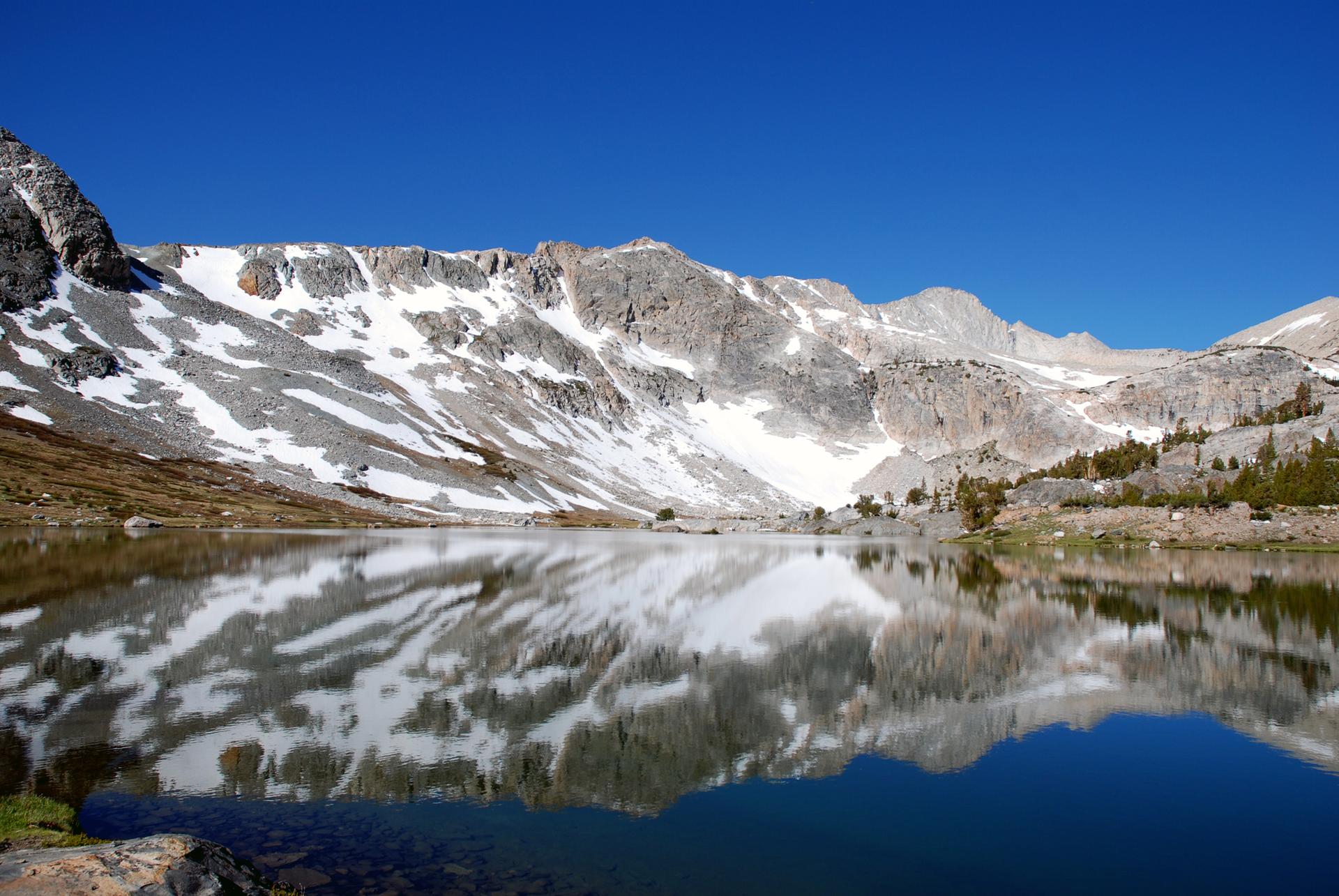 Calm mountain lake reflecting snow-capped peaks under a clear blue sky.