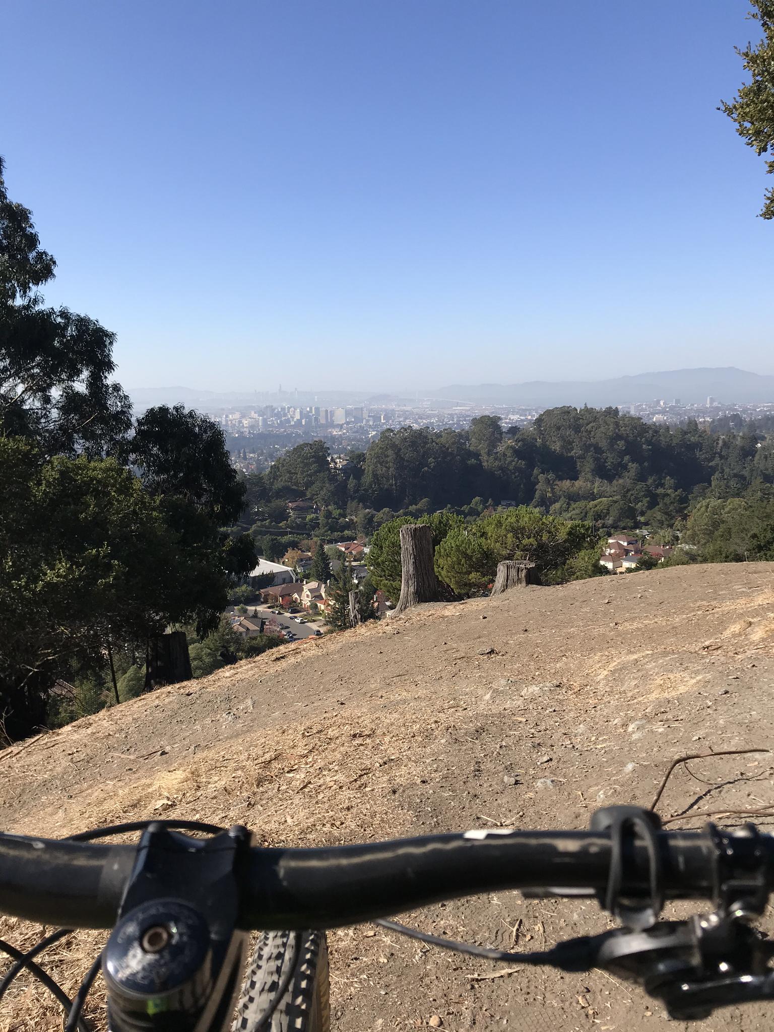 Mountain bike handlebars overlook a distant city and green hills.
