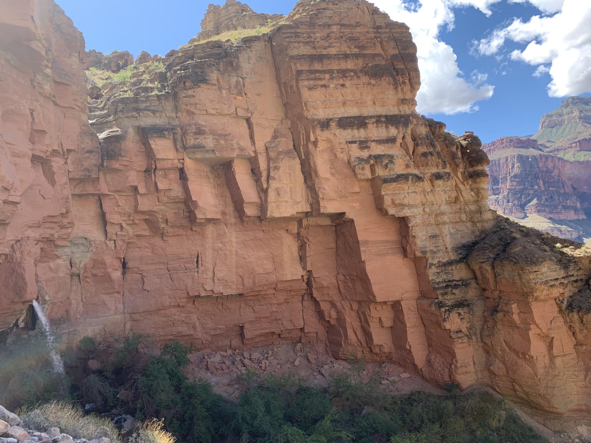 Towering red rock cliff face with green vegetation at its base, under a blue sky.