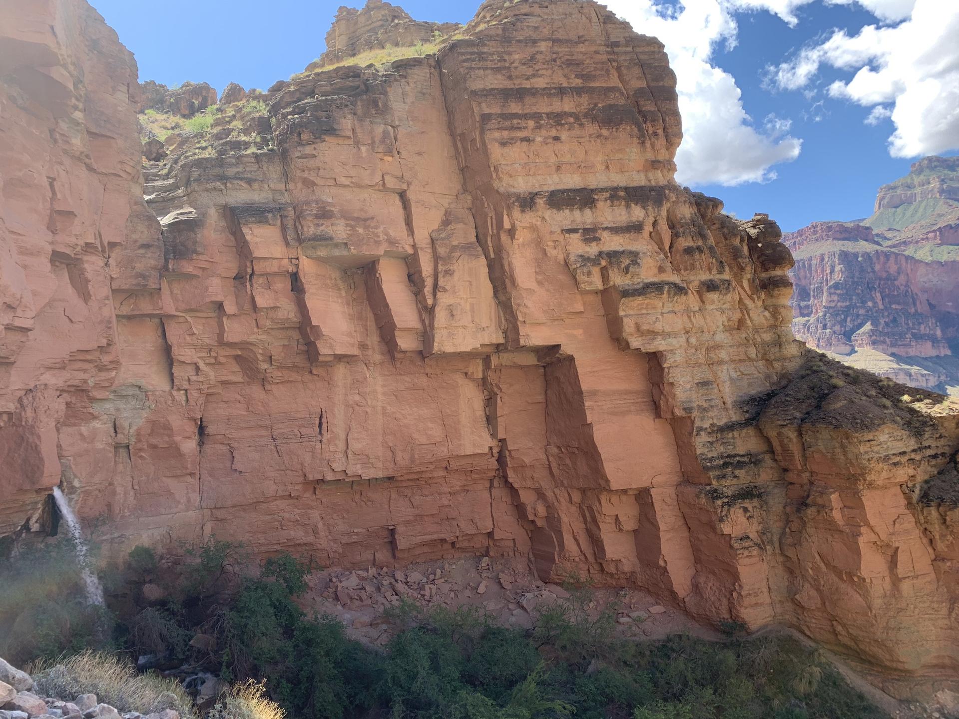 Towering red rock cliff face with green vegetation at its base, under a blue sky.