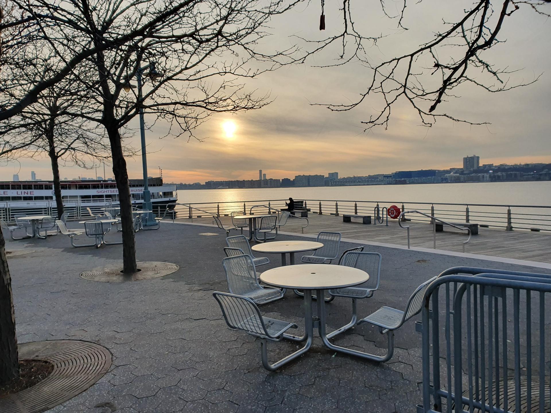 Outdoor tables and chairs on a waterfront promenade at sunset, distant city.