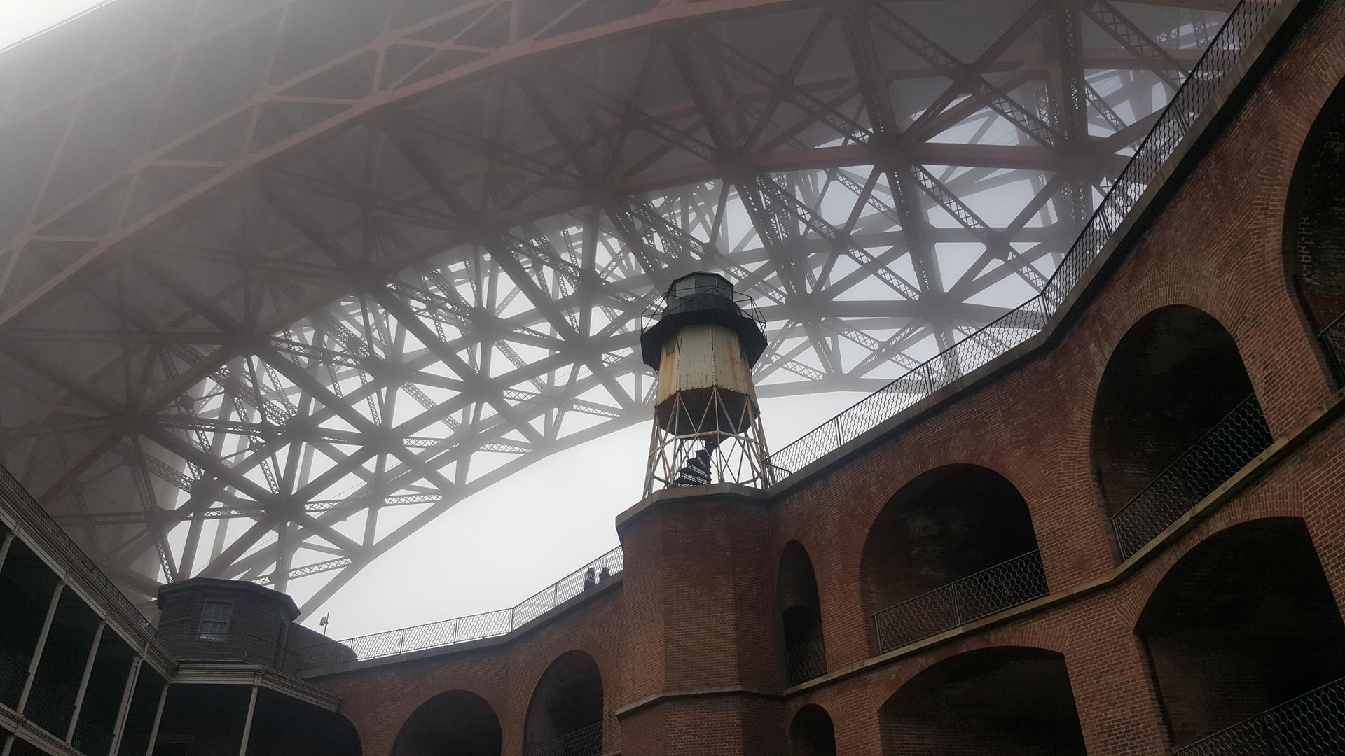 Lighthouse and brick structure under a large bridge in dense fog.