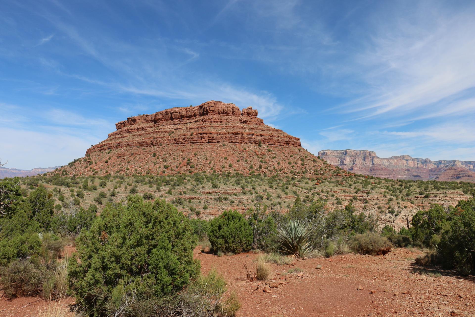Large, flat-topped red rock formation in a sunny desert landscape with scrub.