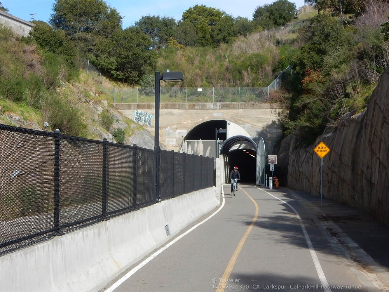 Cyclist rides on a paved path towards a tunnel entrance on a sunny day.