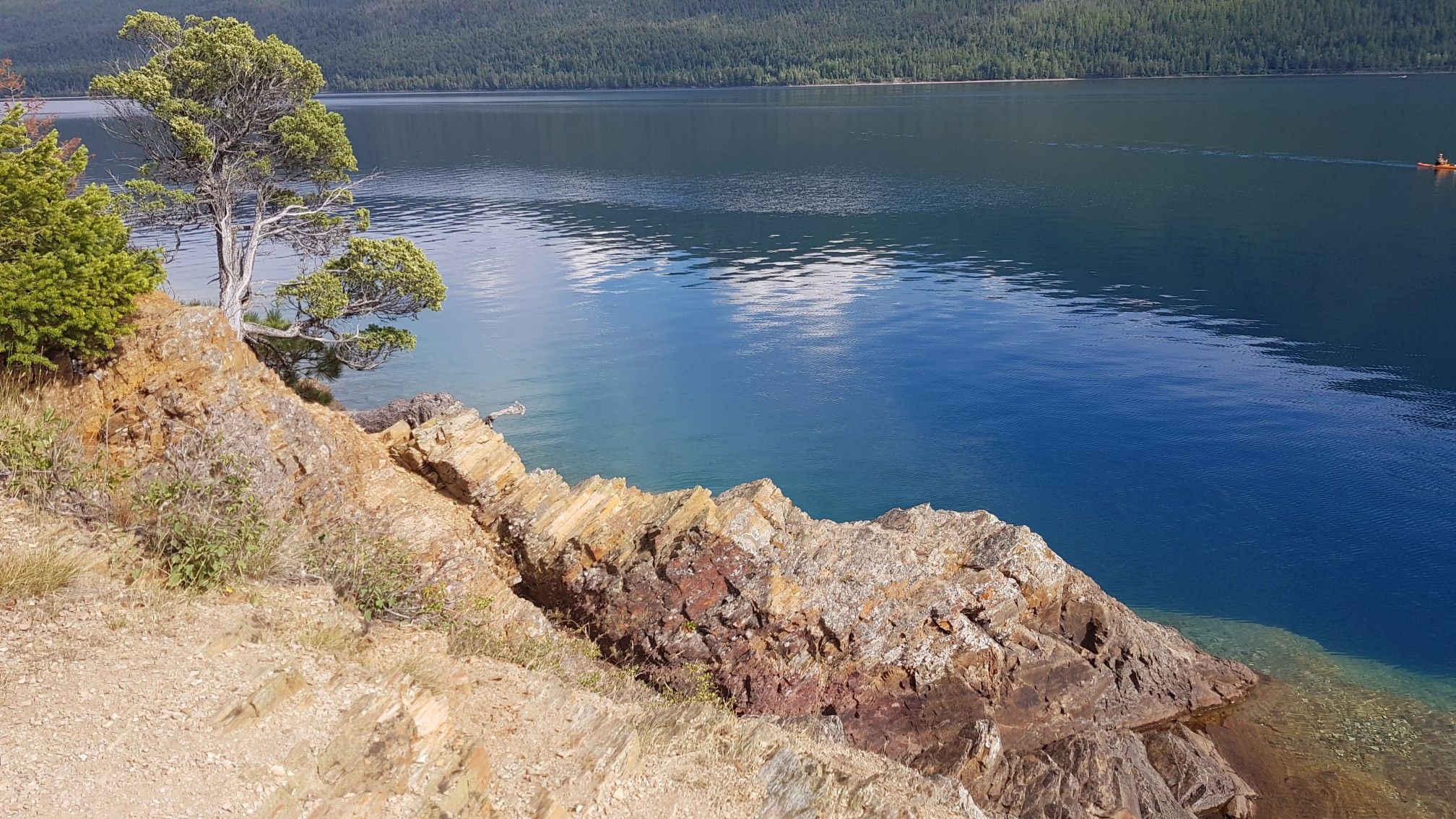 Deep blue lake next to a rocky cliff with distant kayaker and green trees. - Rocky Point Trail