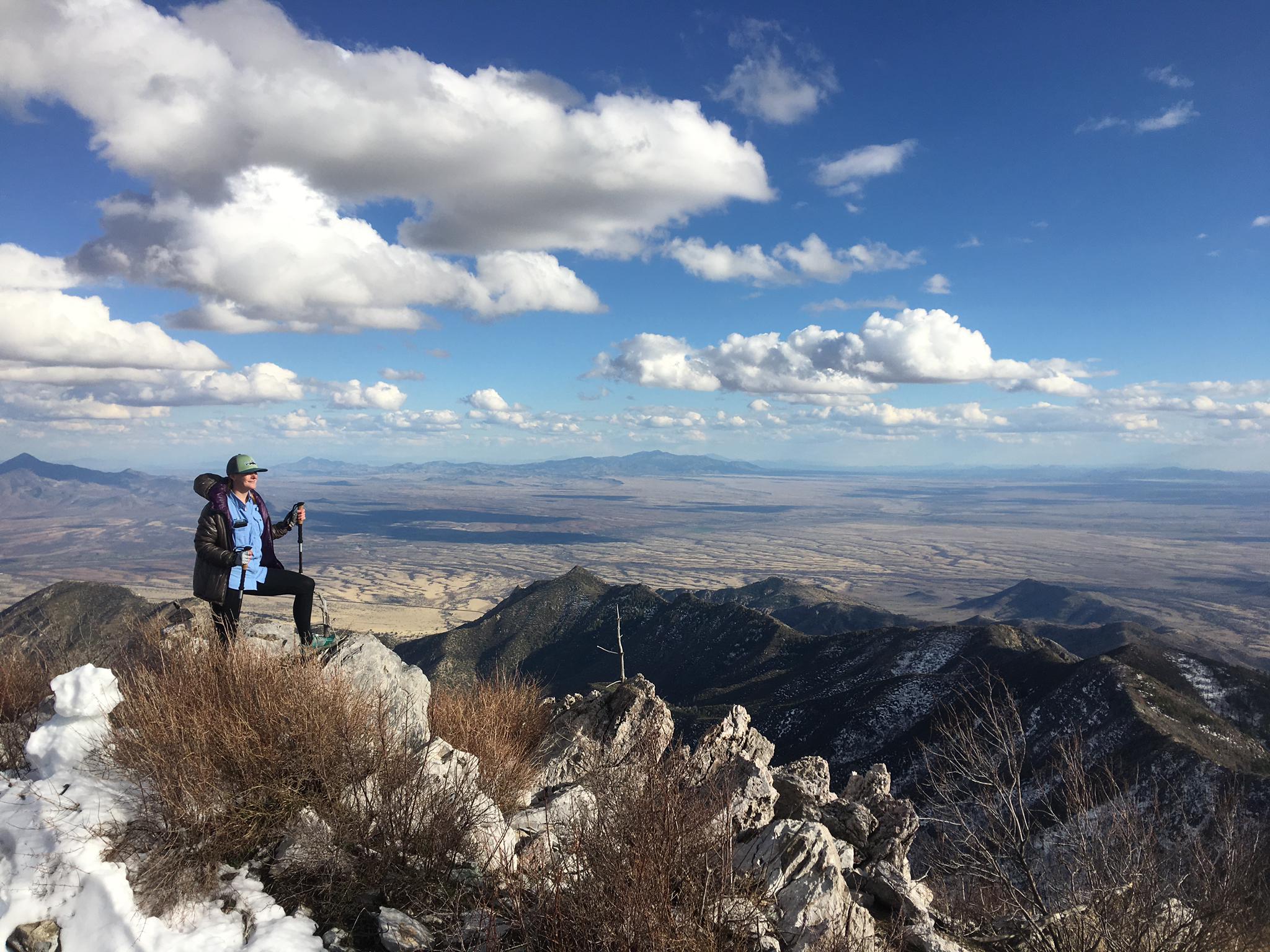 Hiker with trekking pole on a snowy mountain peak overlooking a vast landscape.