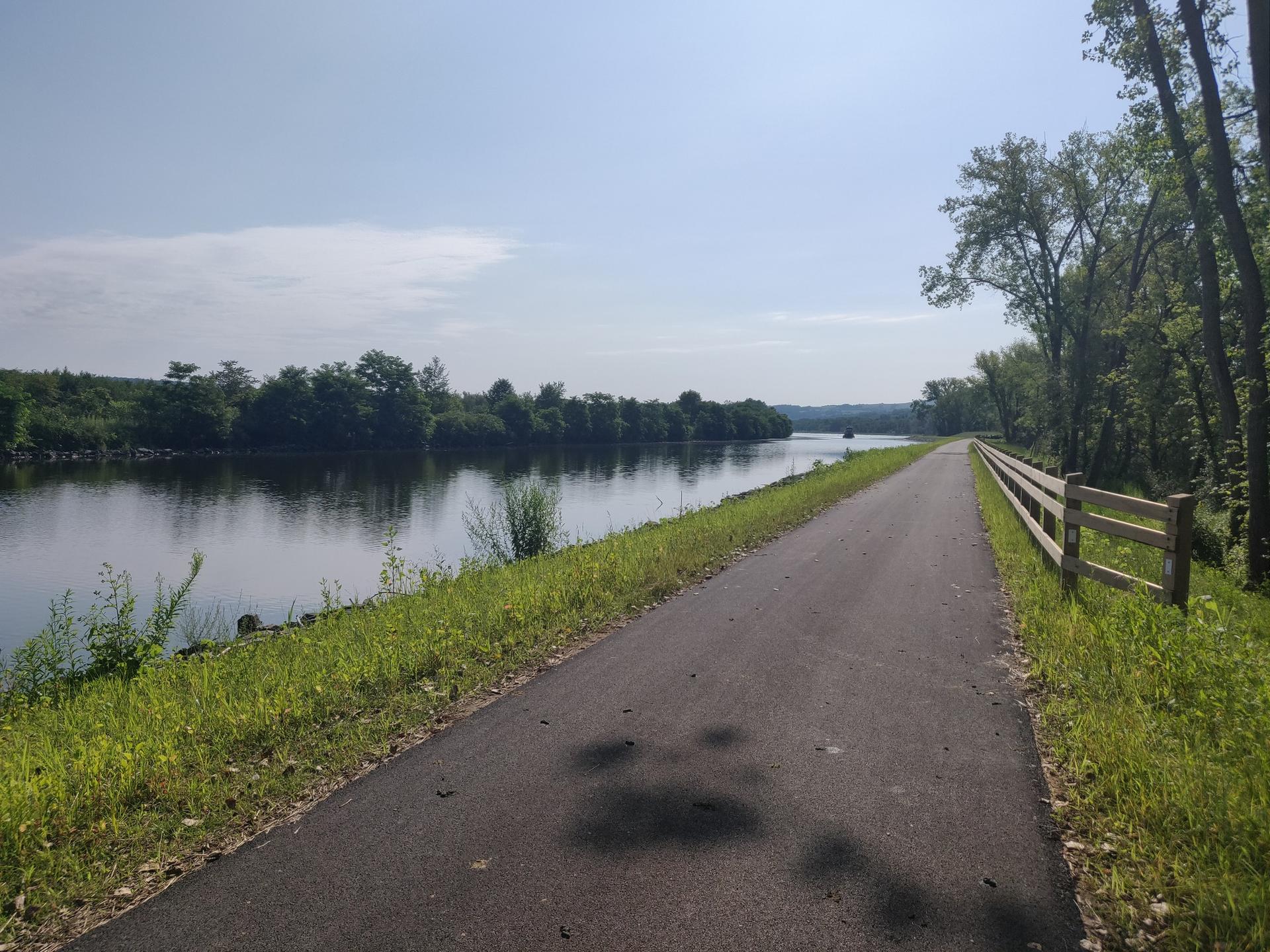 Paved path beside a calm river, green trees, and a wooden fence. Sunny.