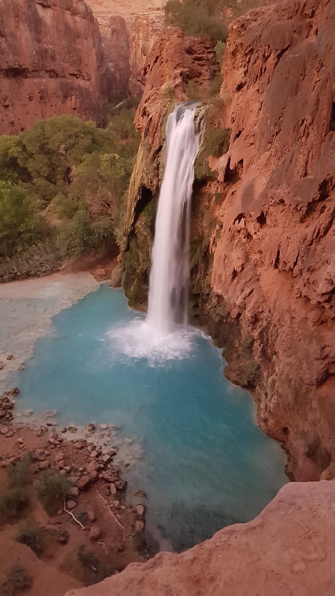 Tall waterfall cascades into a vibrant blue-green pool, framed by red rock.