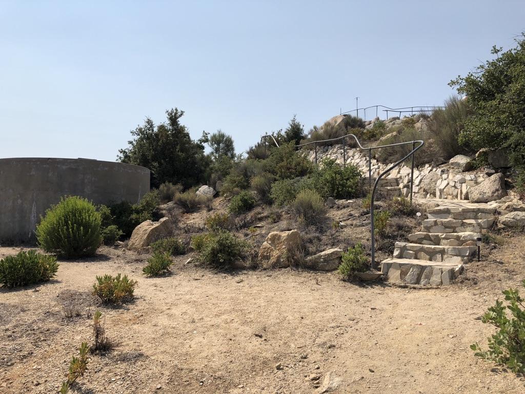 Stone steps with a metal railing lead up a dry, rocky hill under a clear sky.