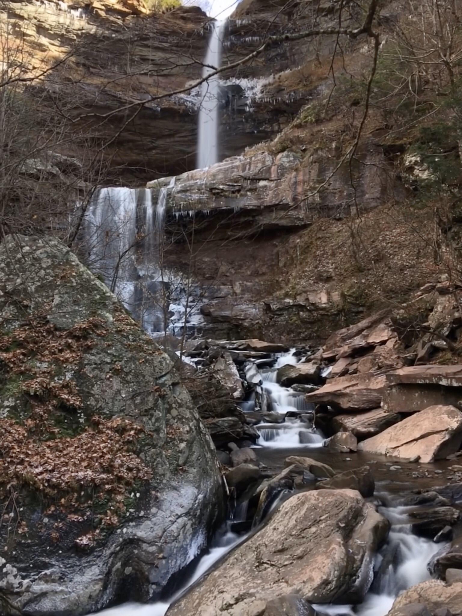 Tall waterfall cascades over rocky cliffs into a stream, surrounded by bare trees.