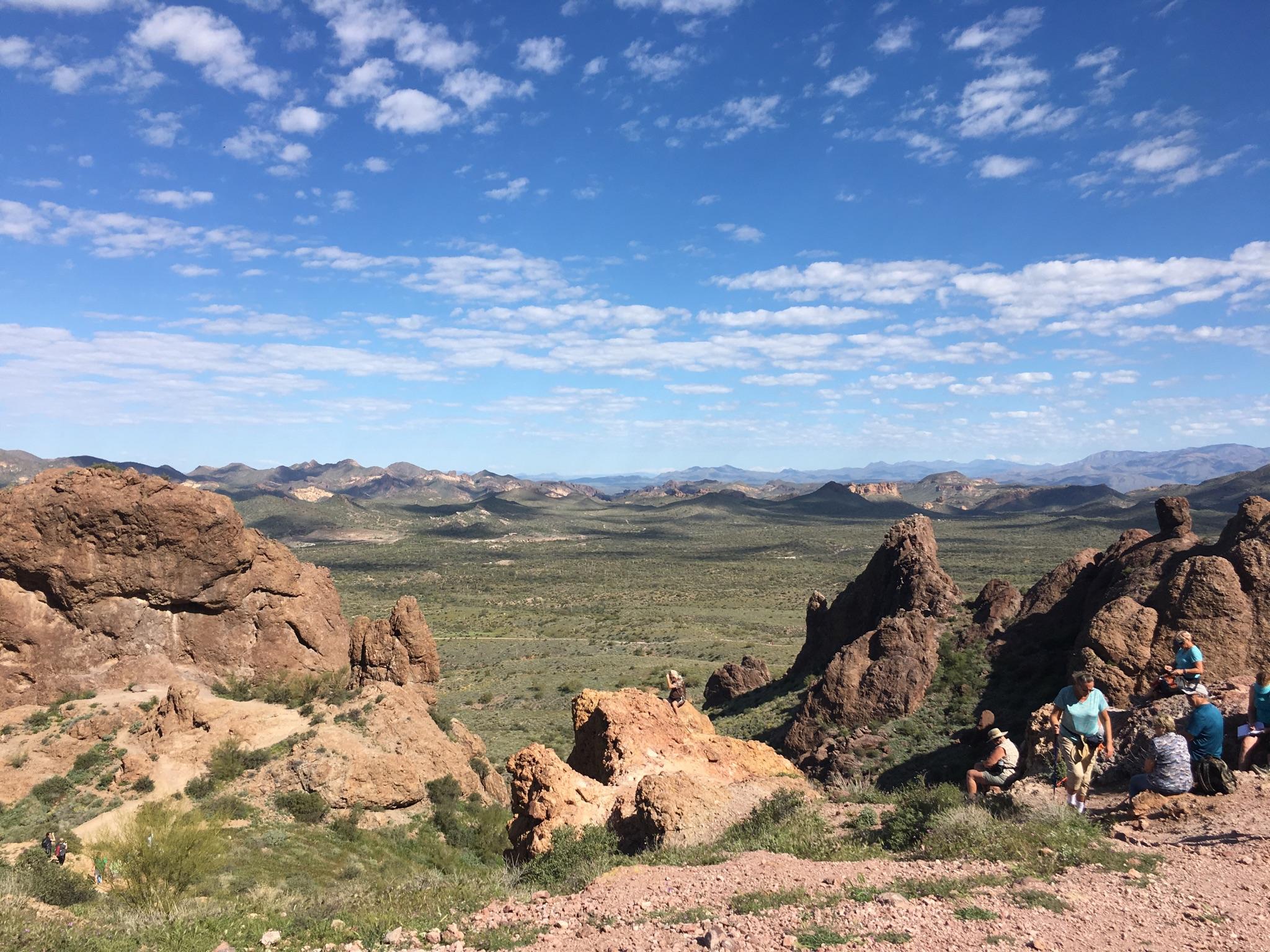 Wide desert landscape with distant mountains, scattered rocks, and people resting.