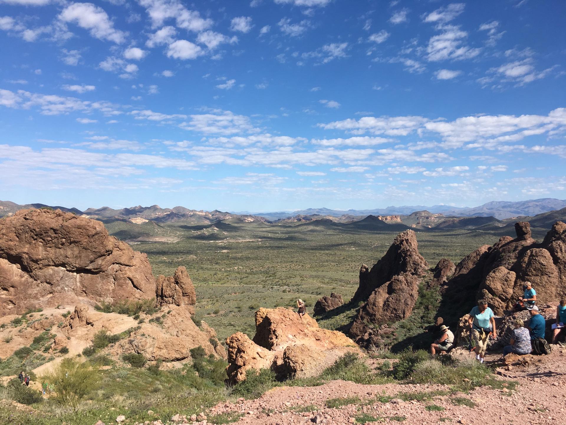 Wide desert landscape with distant mountains, scattered rocks, and people resting.