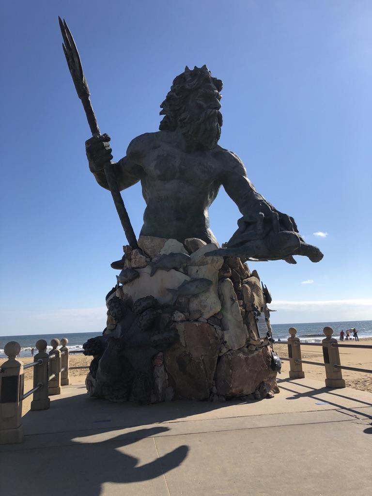 A large, dark statue of a bearded figure holding a trident on a sunny beach.