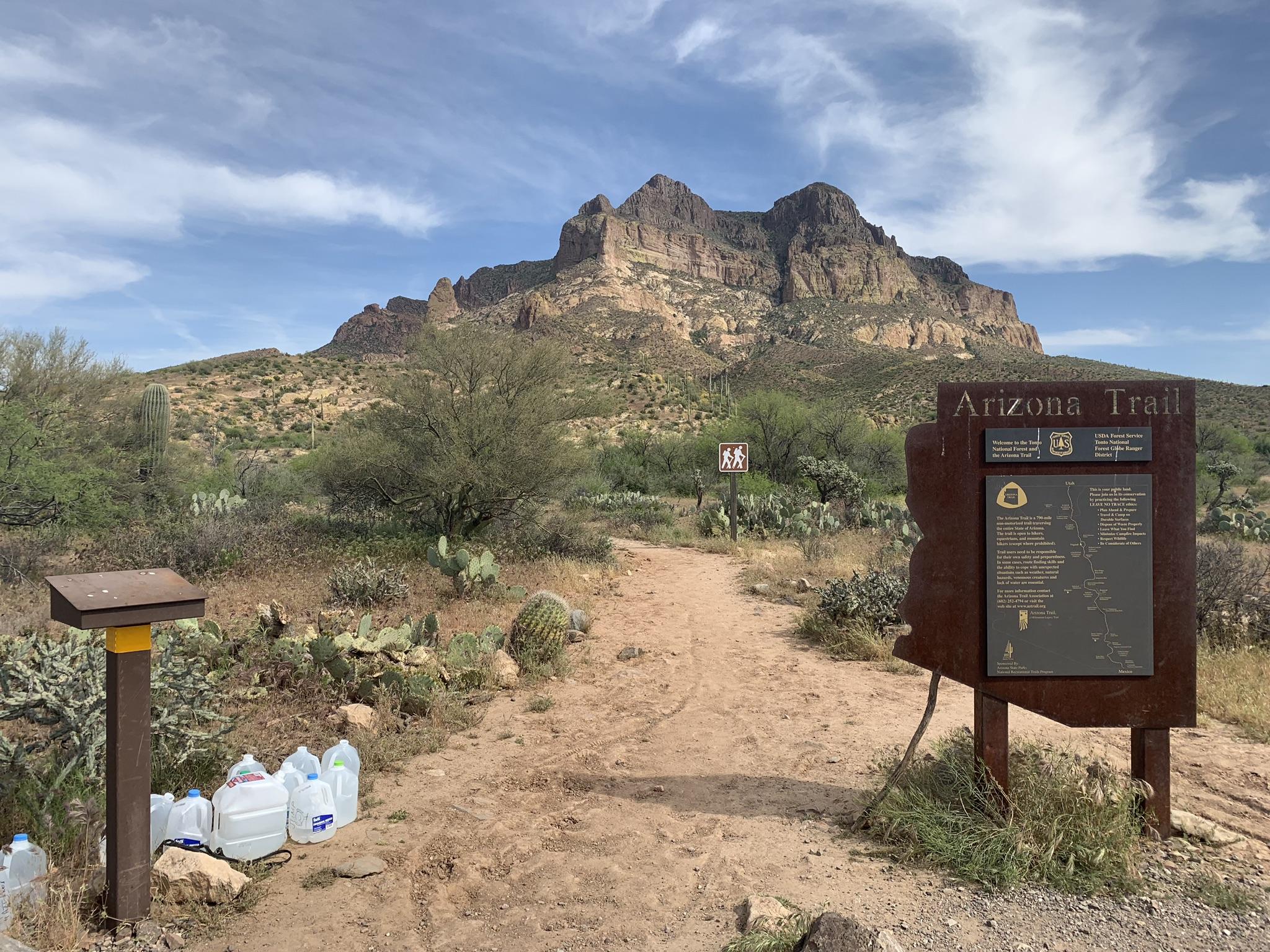 Dirt path leading to a large mountain, with a trail sign and water jugs.