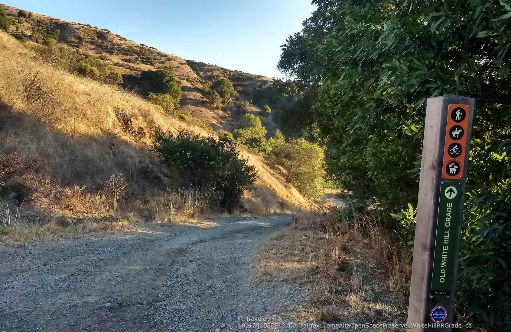Trail marker next to a gravel path, with sunlit hills in the background.