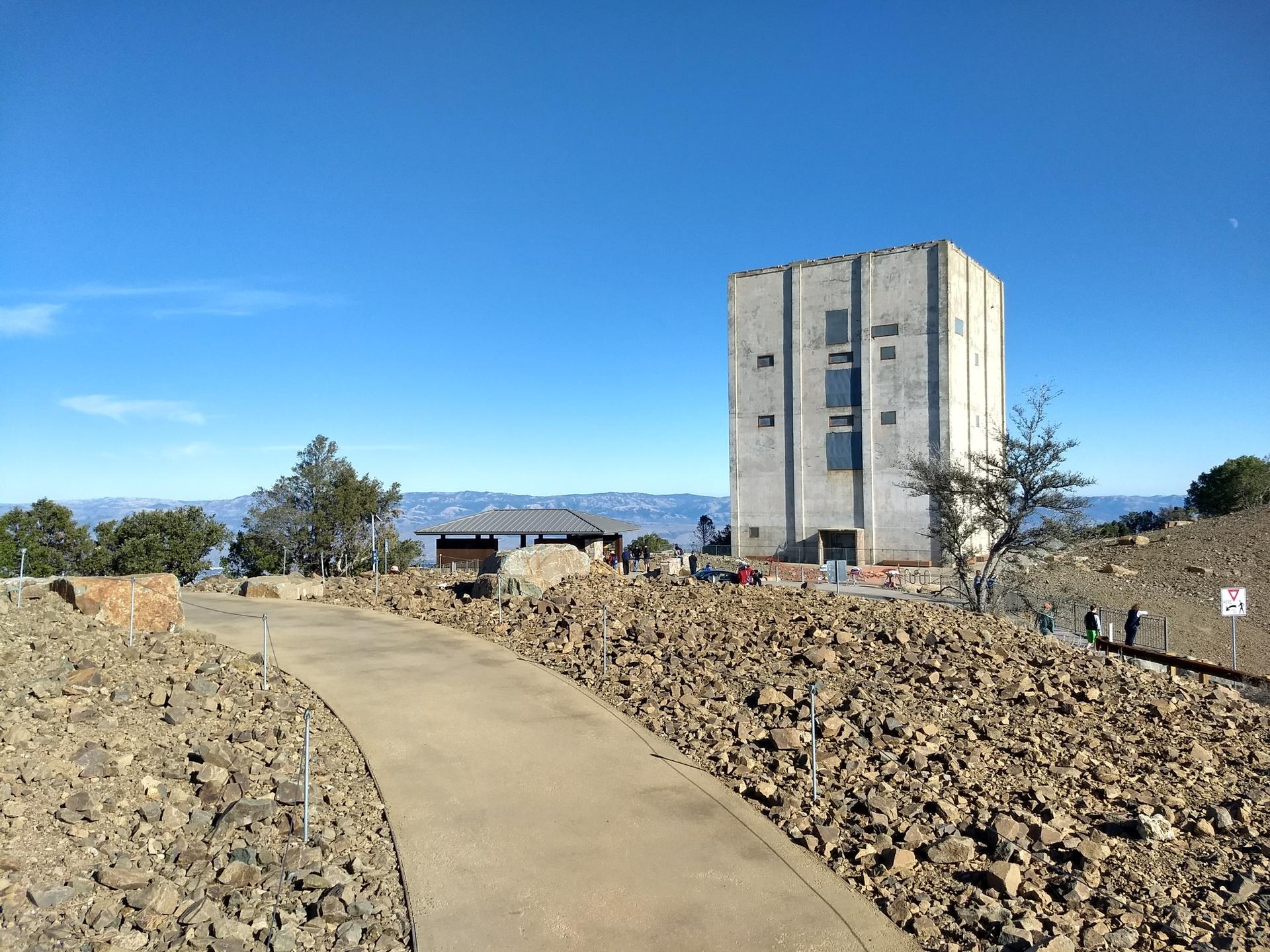 Paved path leading to a large concrete building on a rocky hilltop, people visible.