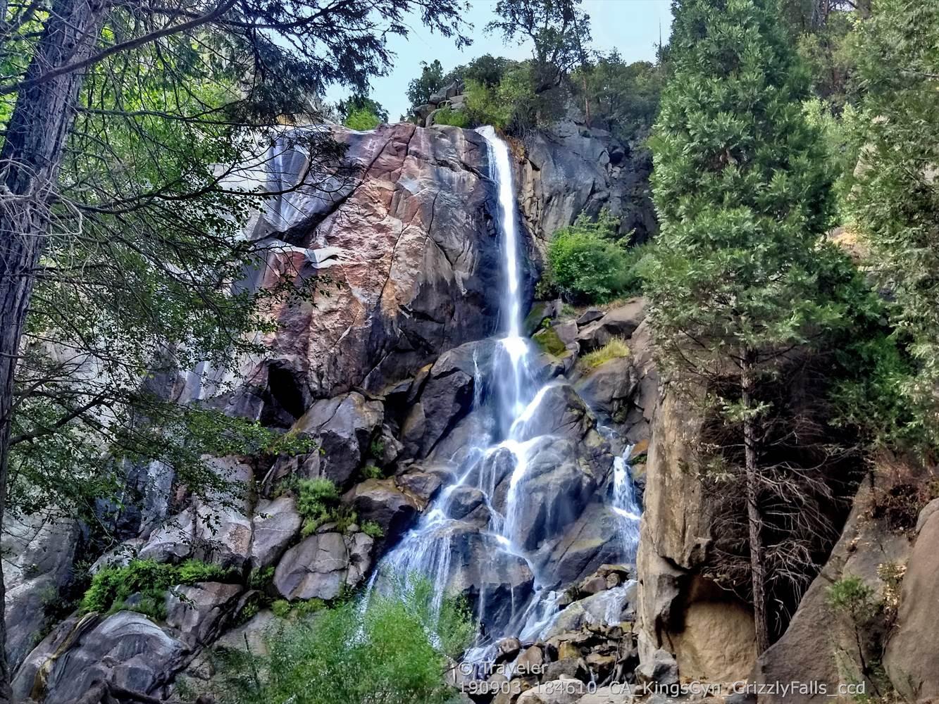 A tall waterfall cascades down a large, rocky cliff surrounded by green trees.