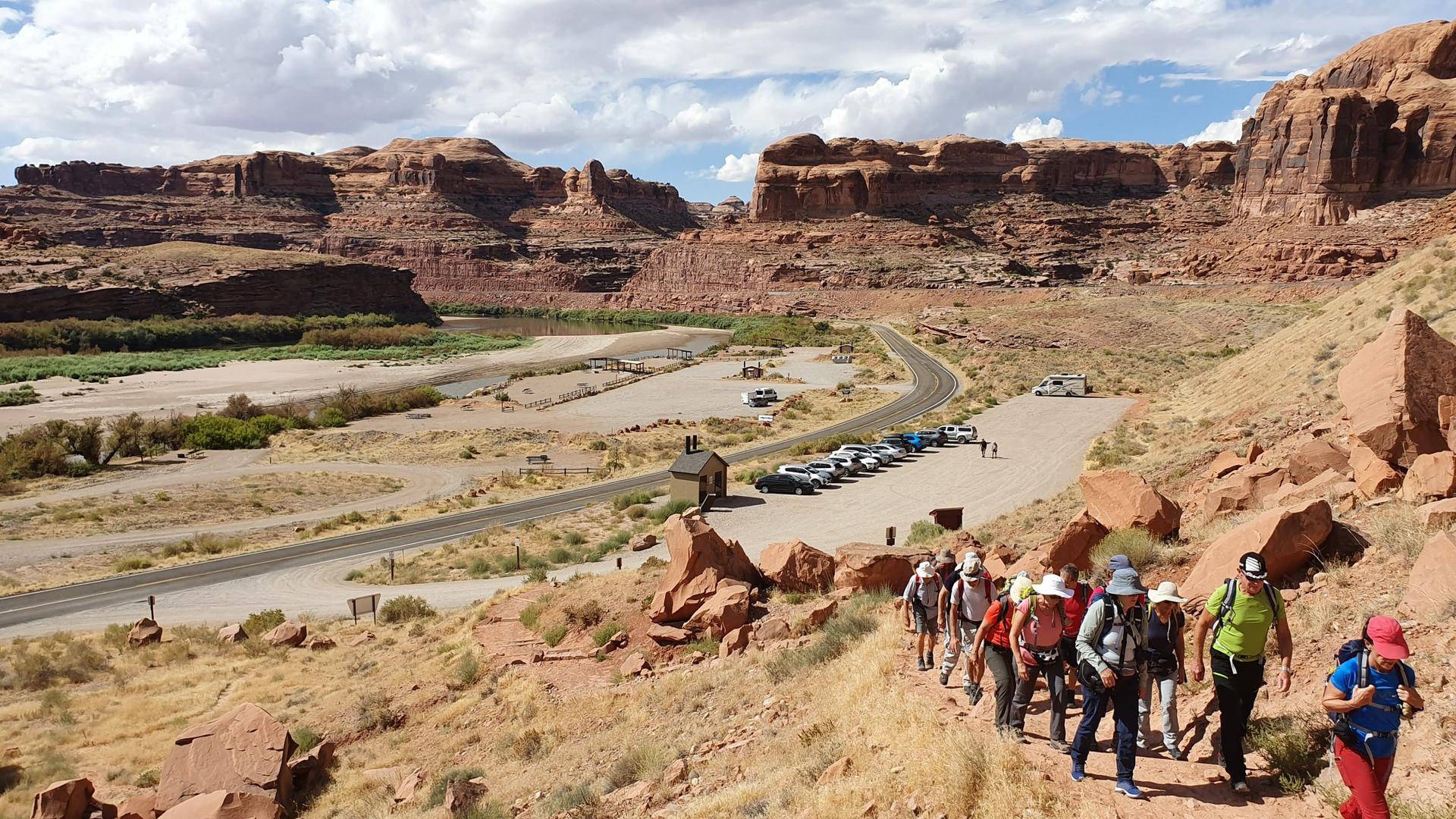 Group of hikers walks along a desert trail overlooking a river and parking area.