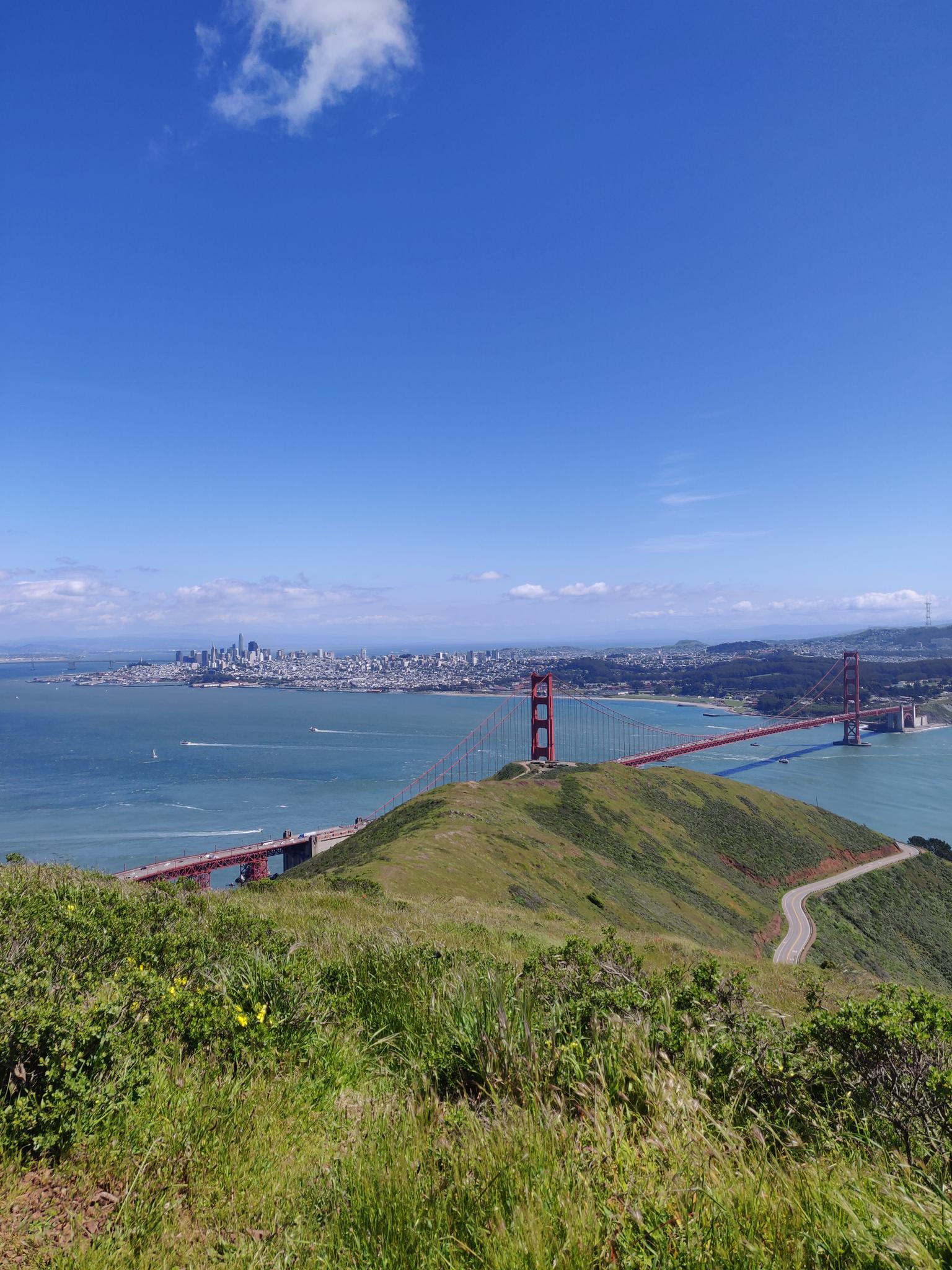 A red suspension bridge spans water, with a distant city skyline under blue sky.