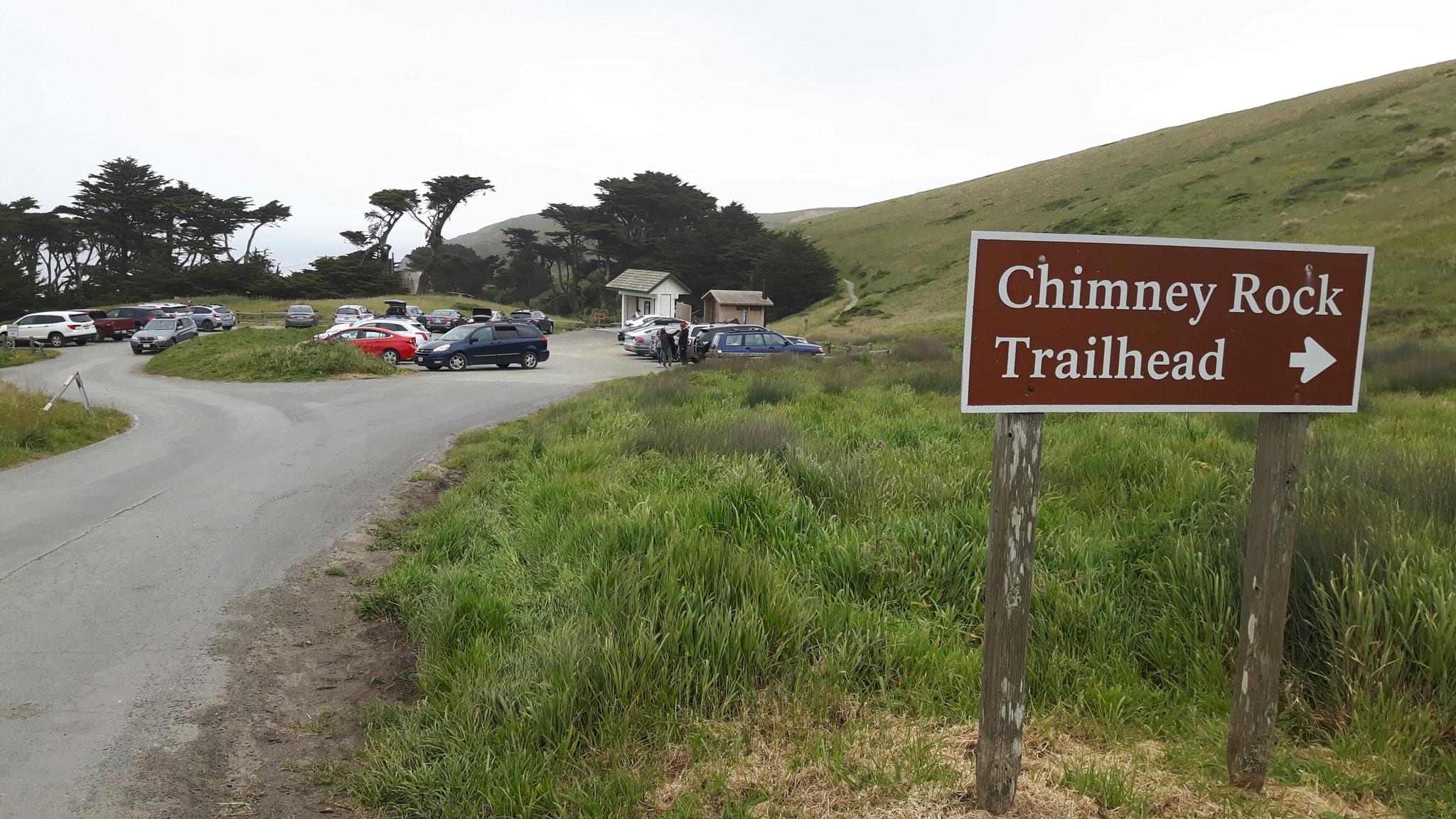 Trailhead sign pointing right, with a parking lot and grassy hills under an overcast sky.