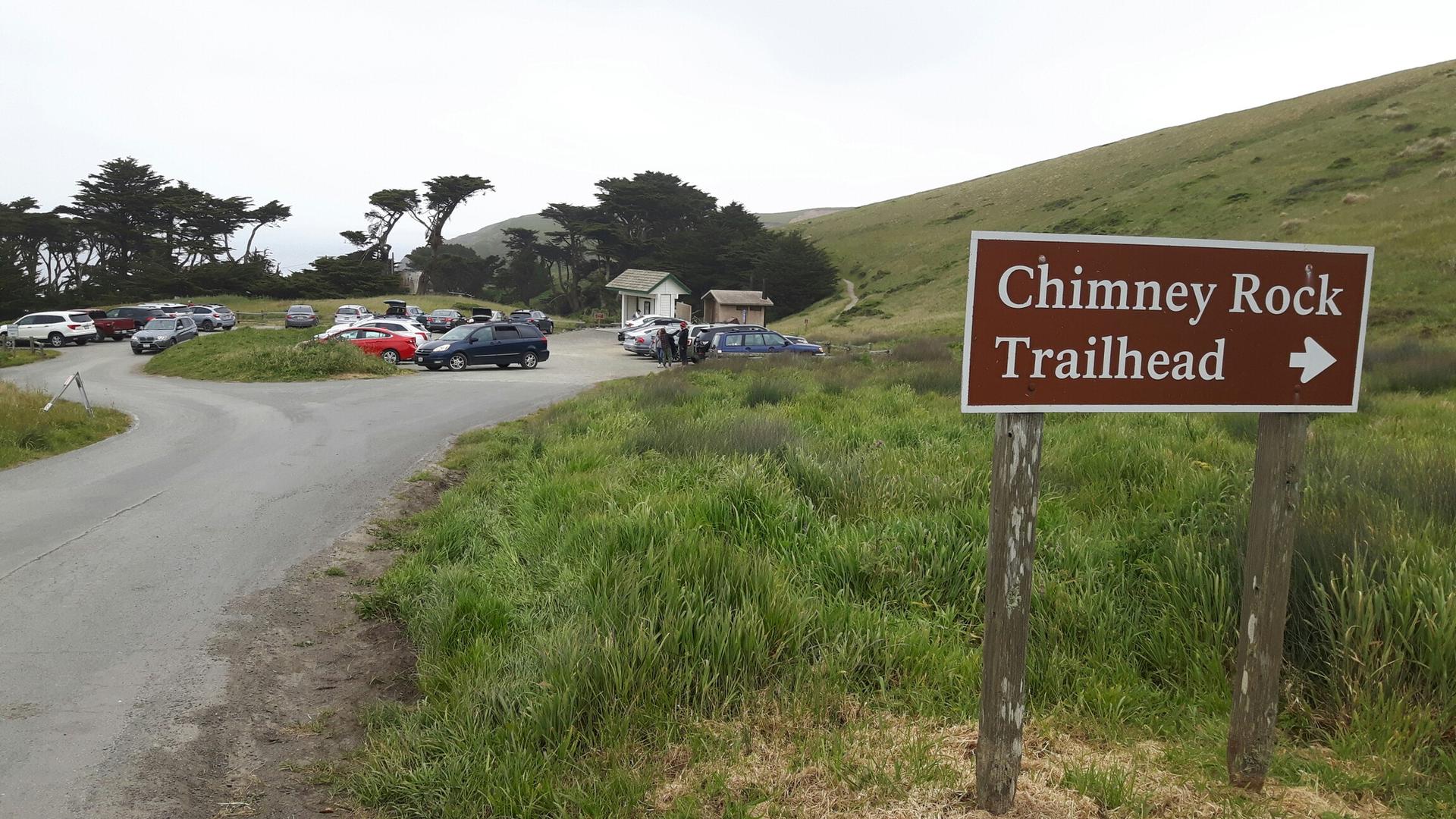Trailhead sign pointing right, with a parking lot and grassy hills under an overcast sky.