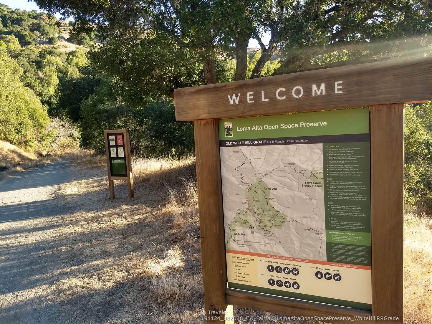 Wooden welcome sign and map on a dirt path, surrounded by trees.