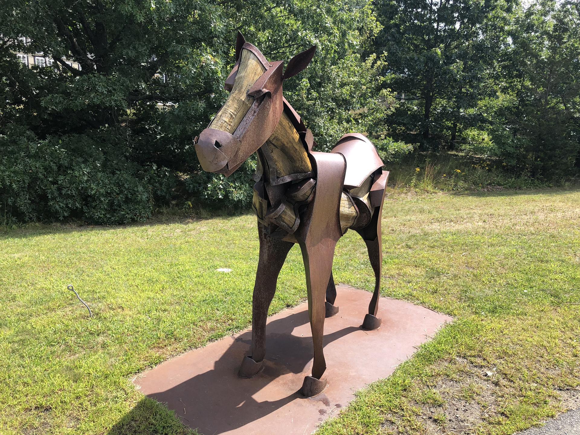 A rustic metal horse sculpture stands on grass under bright sunlight.