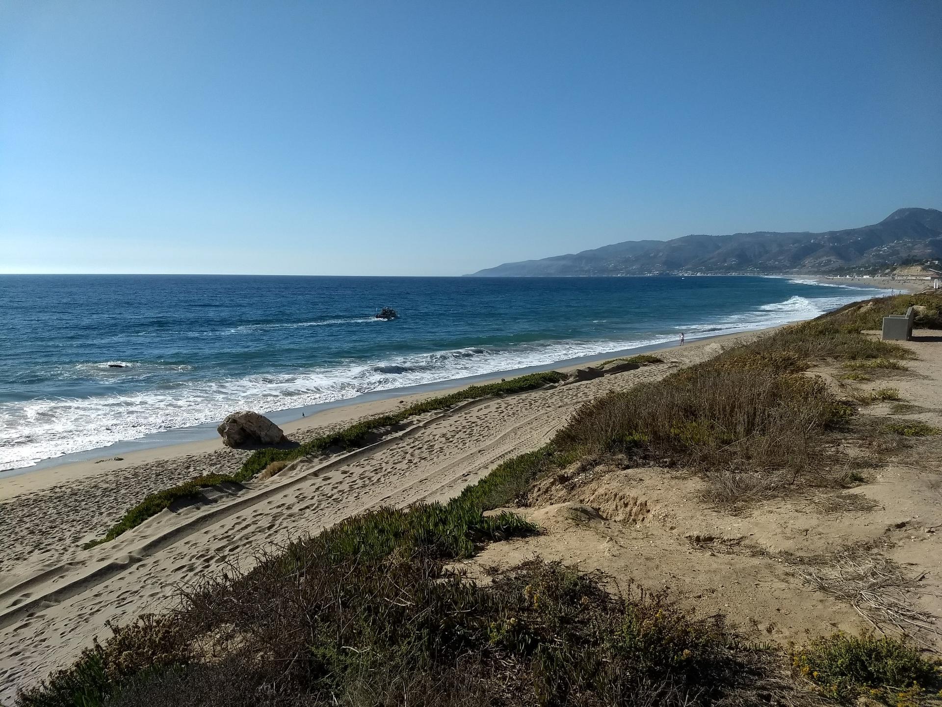 Sunny beach with ocean waves, distant mountains, and a boat in the water.