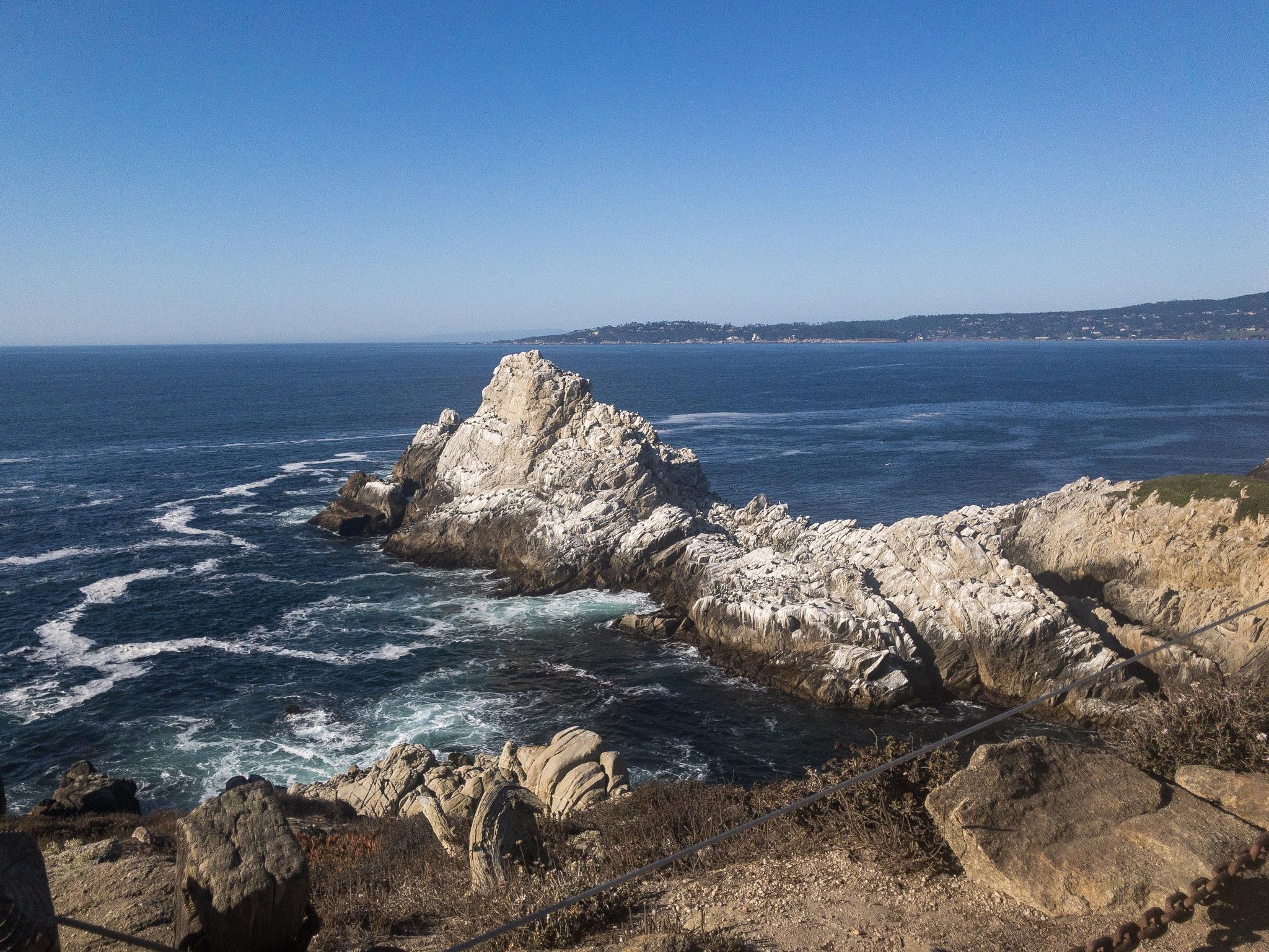 Large white rock formations in the dark blue ocean with distant land under clear sky.