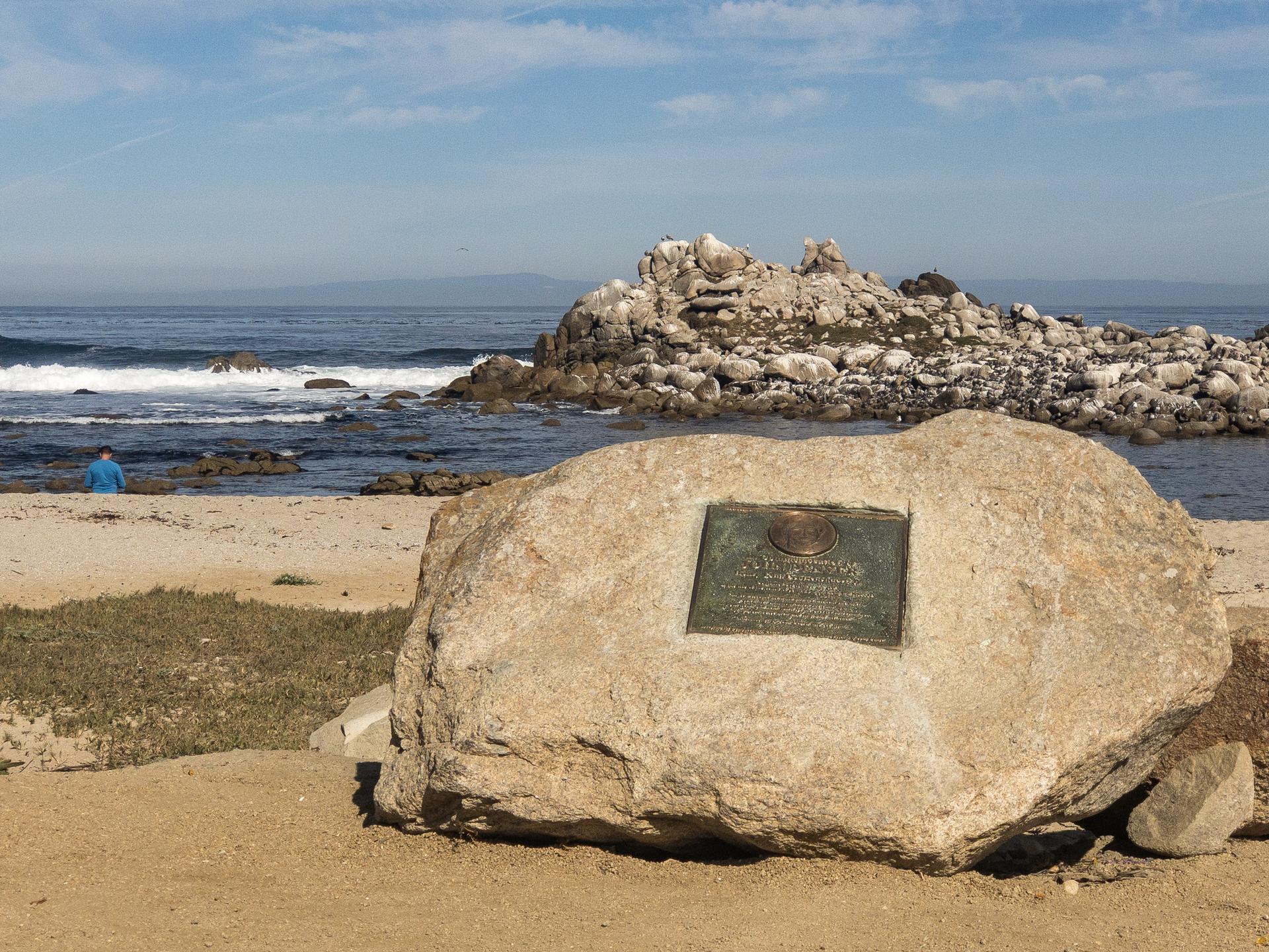 Large rock with plaque on a sandy beach, person by ocean, sunny day.