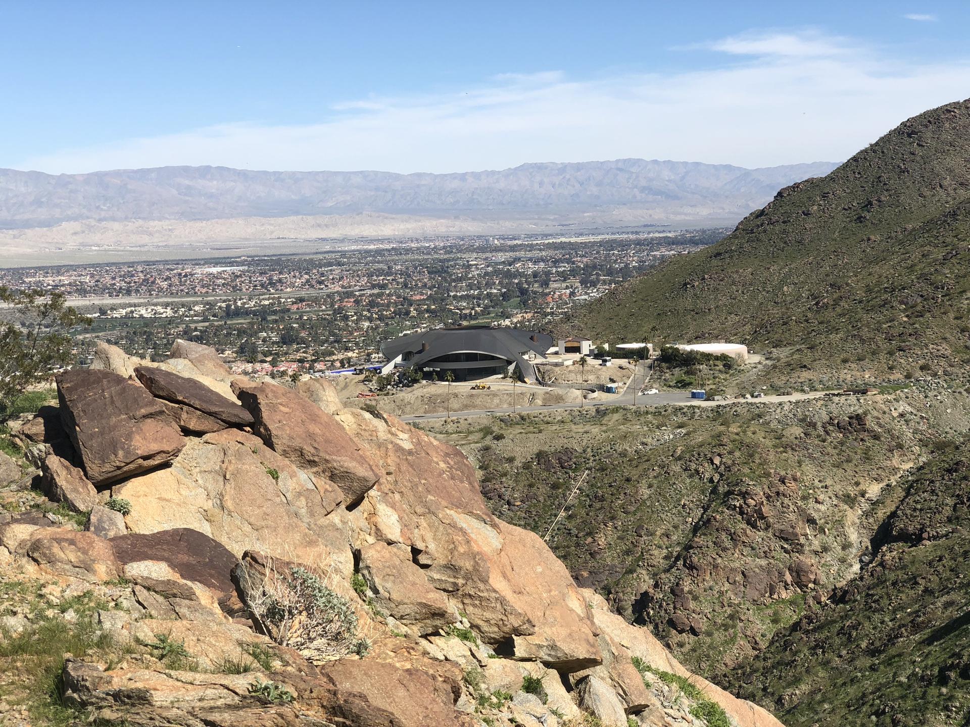 Rocky desert hillside with a modern building and distant city under clear skies.