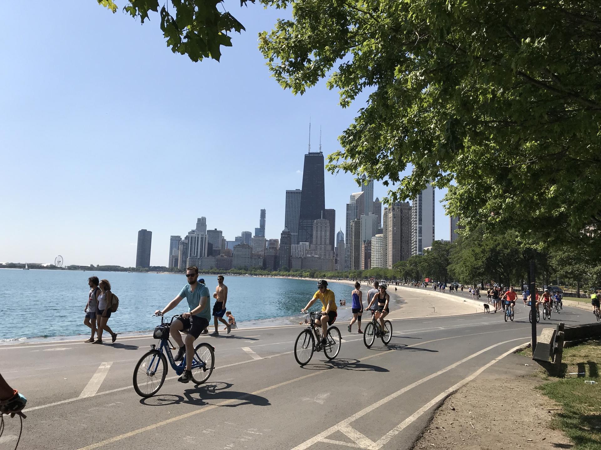 People cycling and walking along a sunny waterfront path with a city skyline.