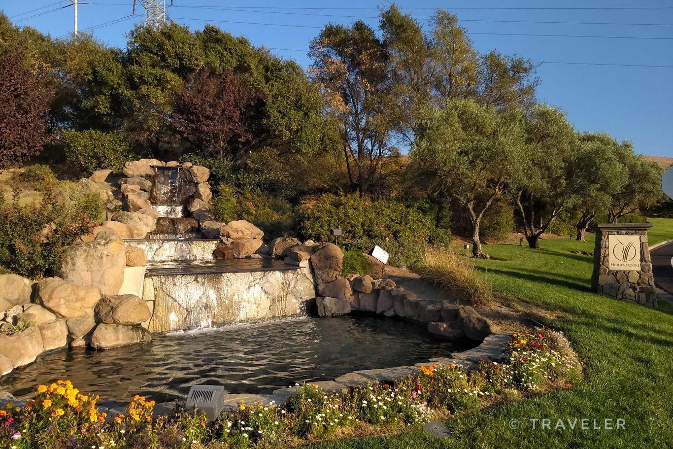 Man-made rock waterfall flowing into a pond, surrounded by green grass.