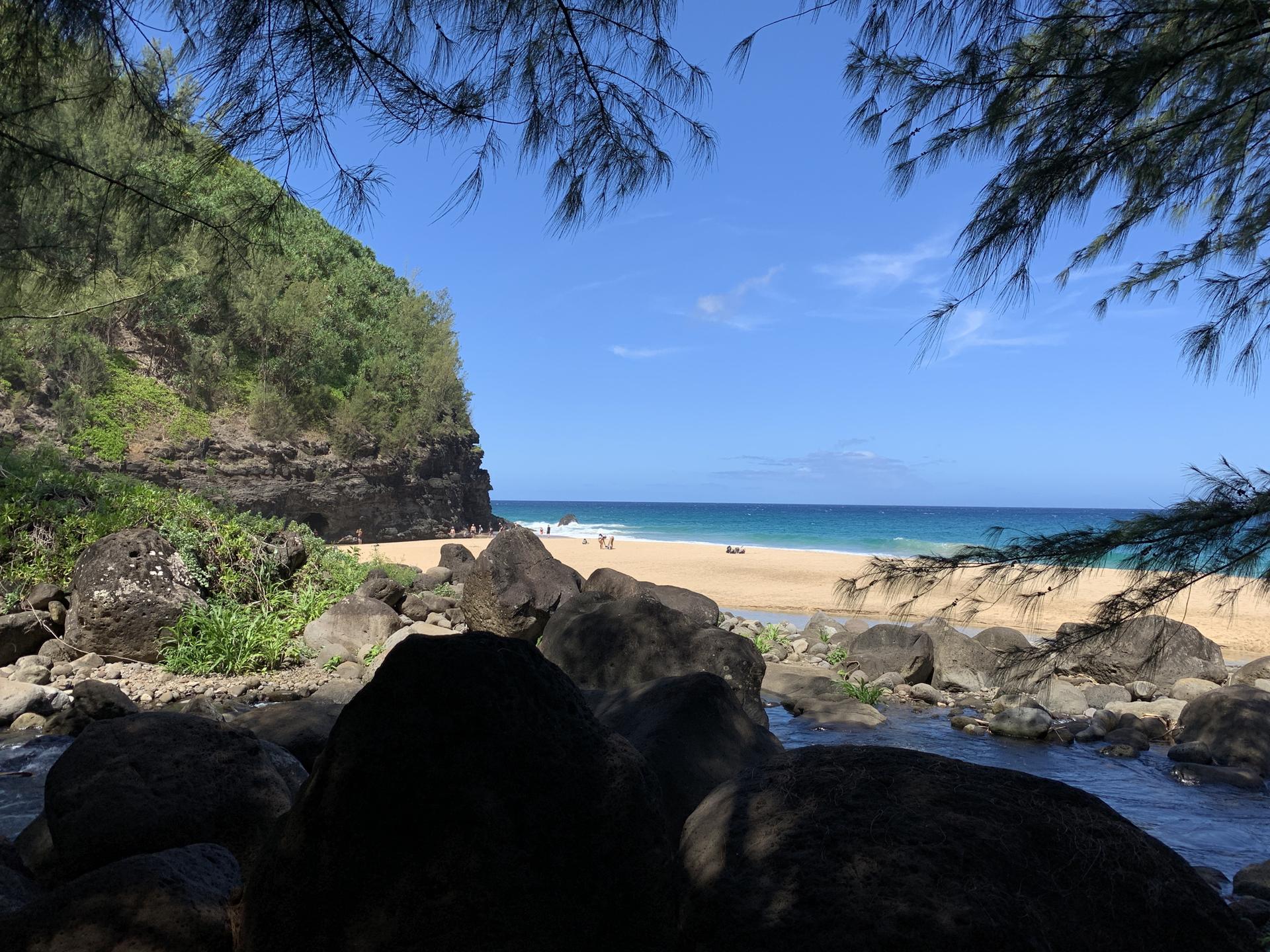 Rocky foreground leads to a sandy beach, blue ocean, and green cliff.