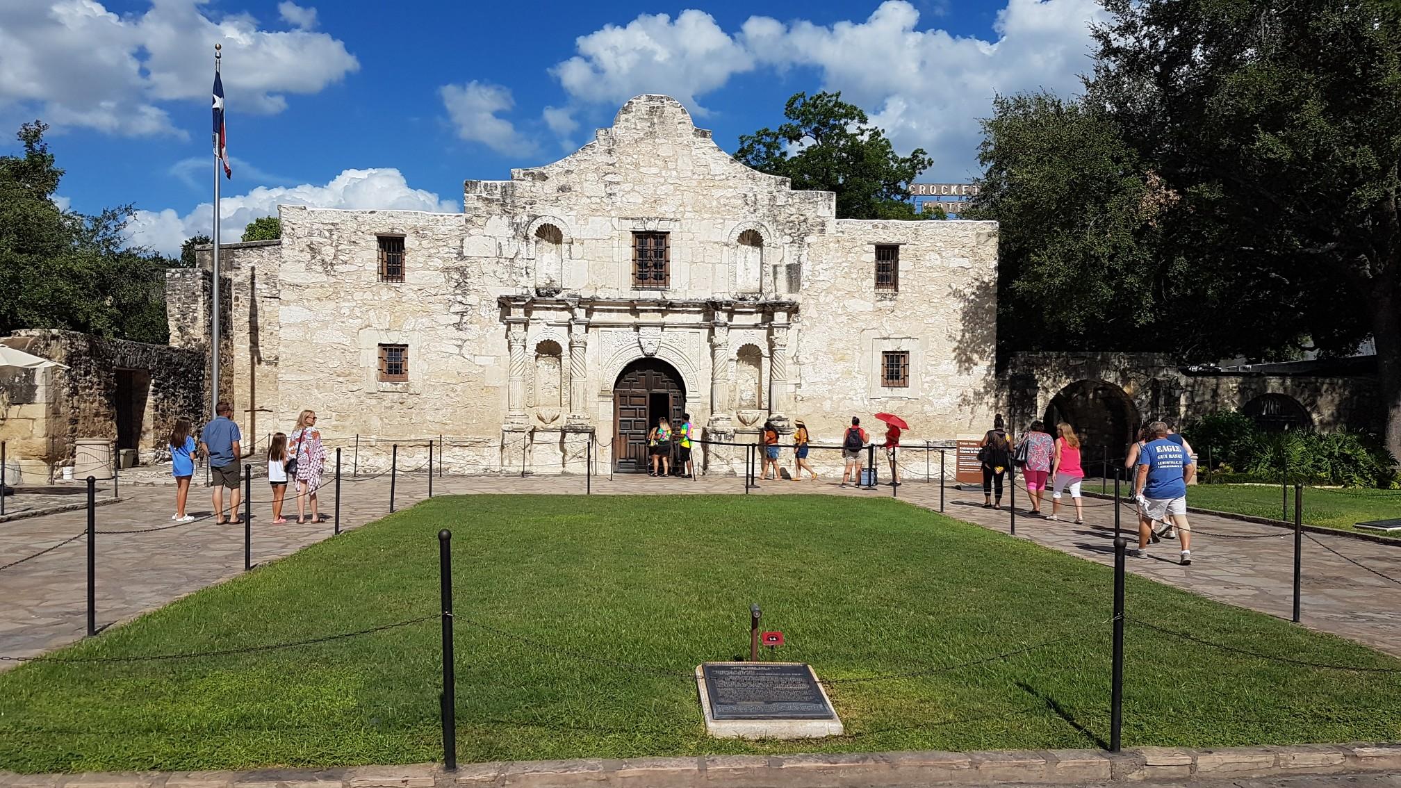 Historic stone building with a flag, people walking on a sunny day.