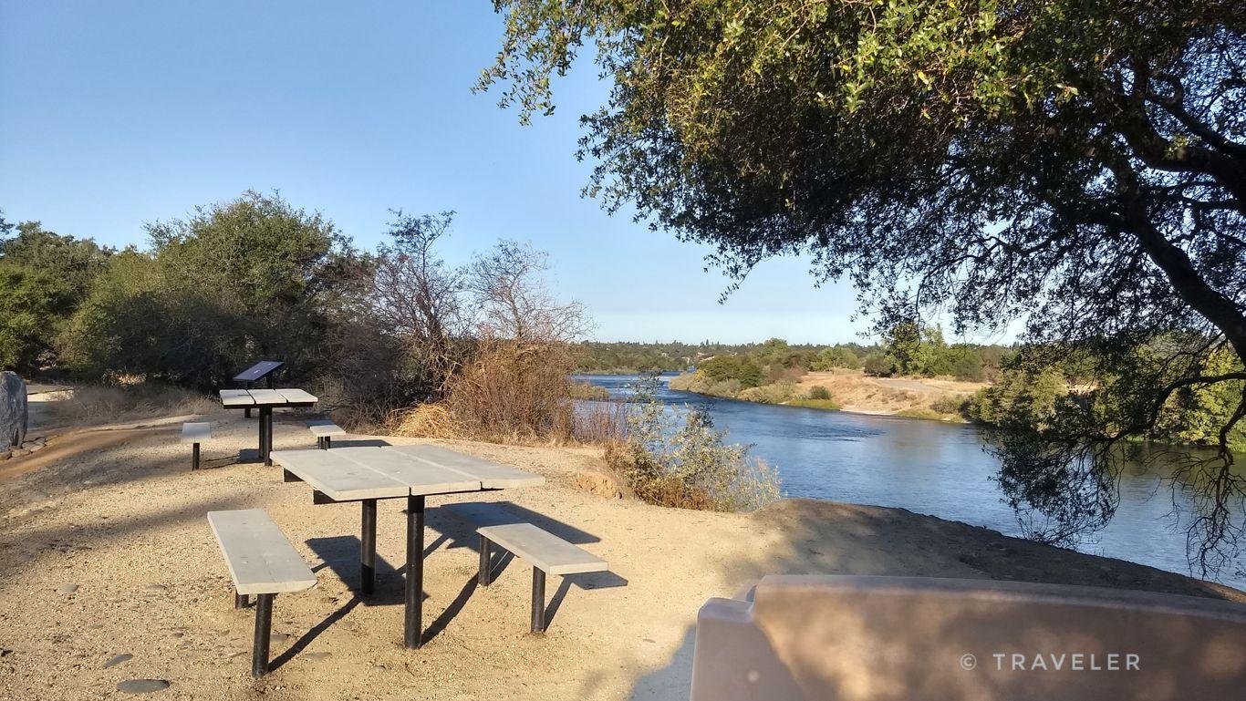 Two picnic tables on a dirt bank overlooking a wide, sunny river.