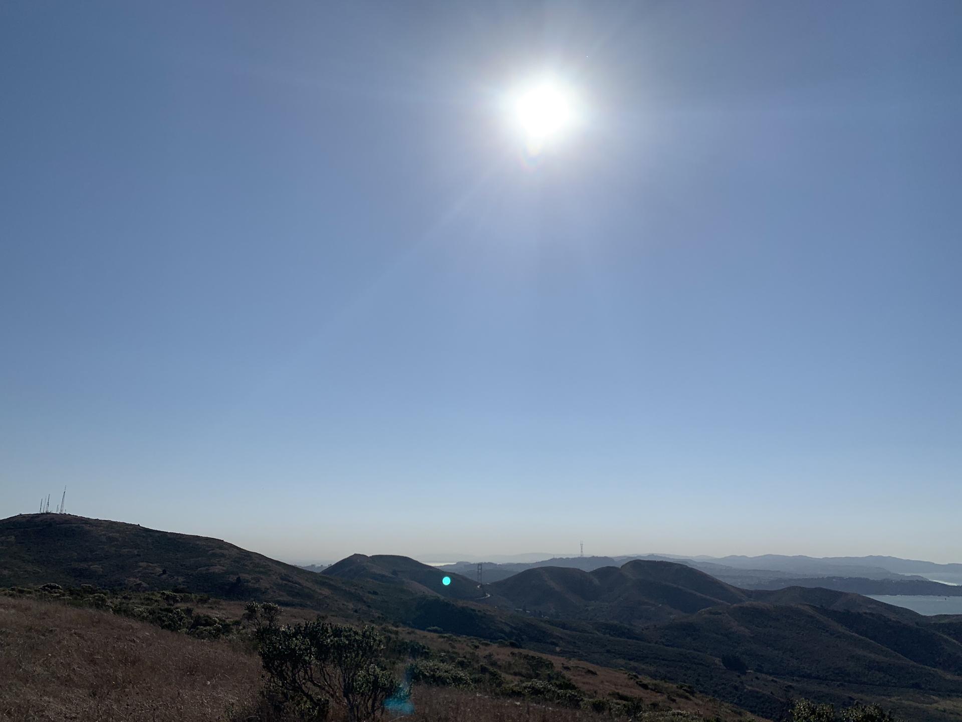 Bright sun in a clear blue sky over rolling, arid hills and distant water.