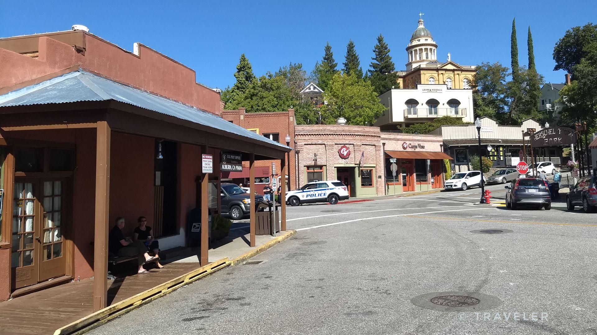 Sunny street scene with historic buildings, parked cars, and people on a porch.