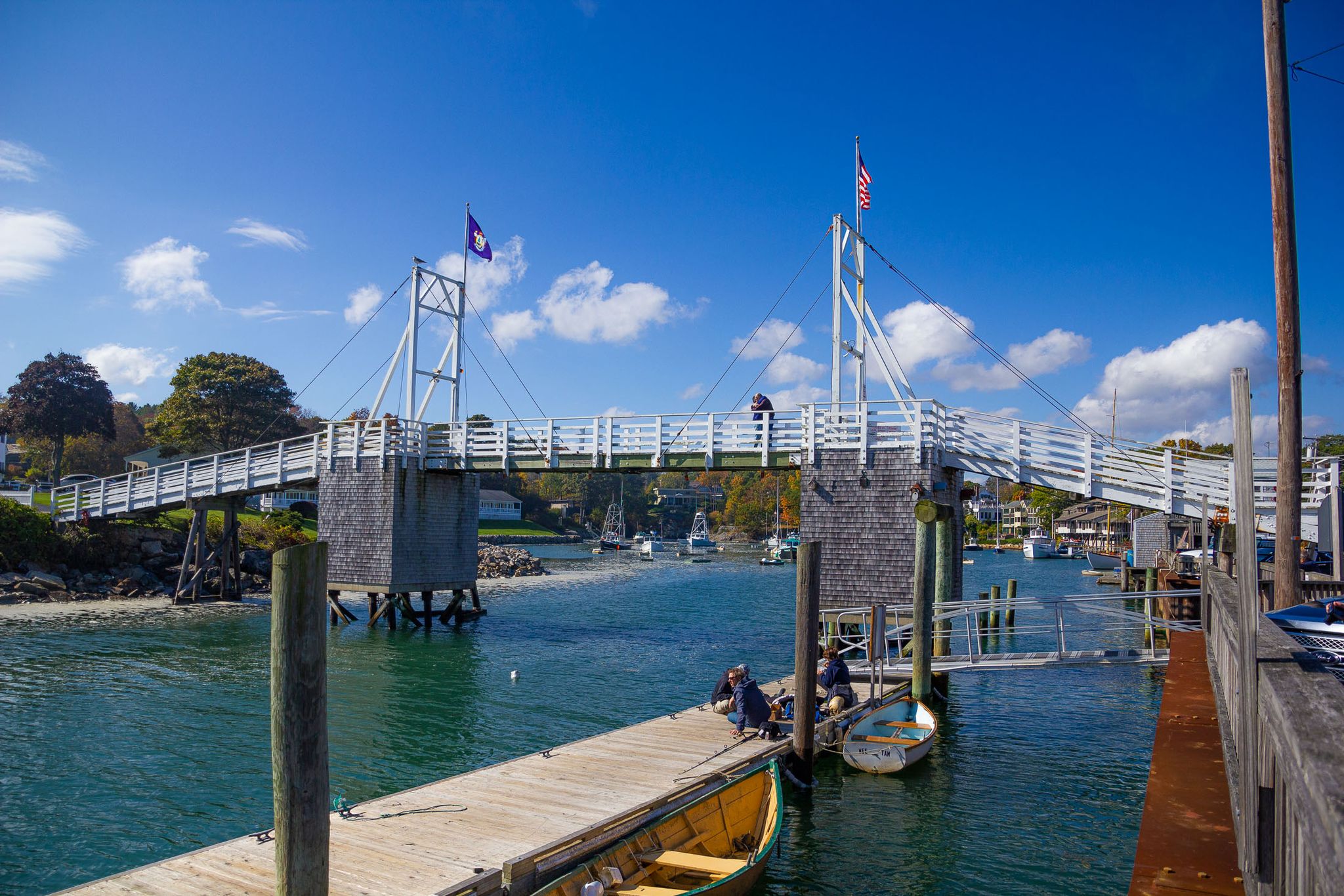 White pedestrian bridge over a calm waterway with boats and people. - Marginal Way
