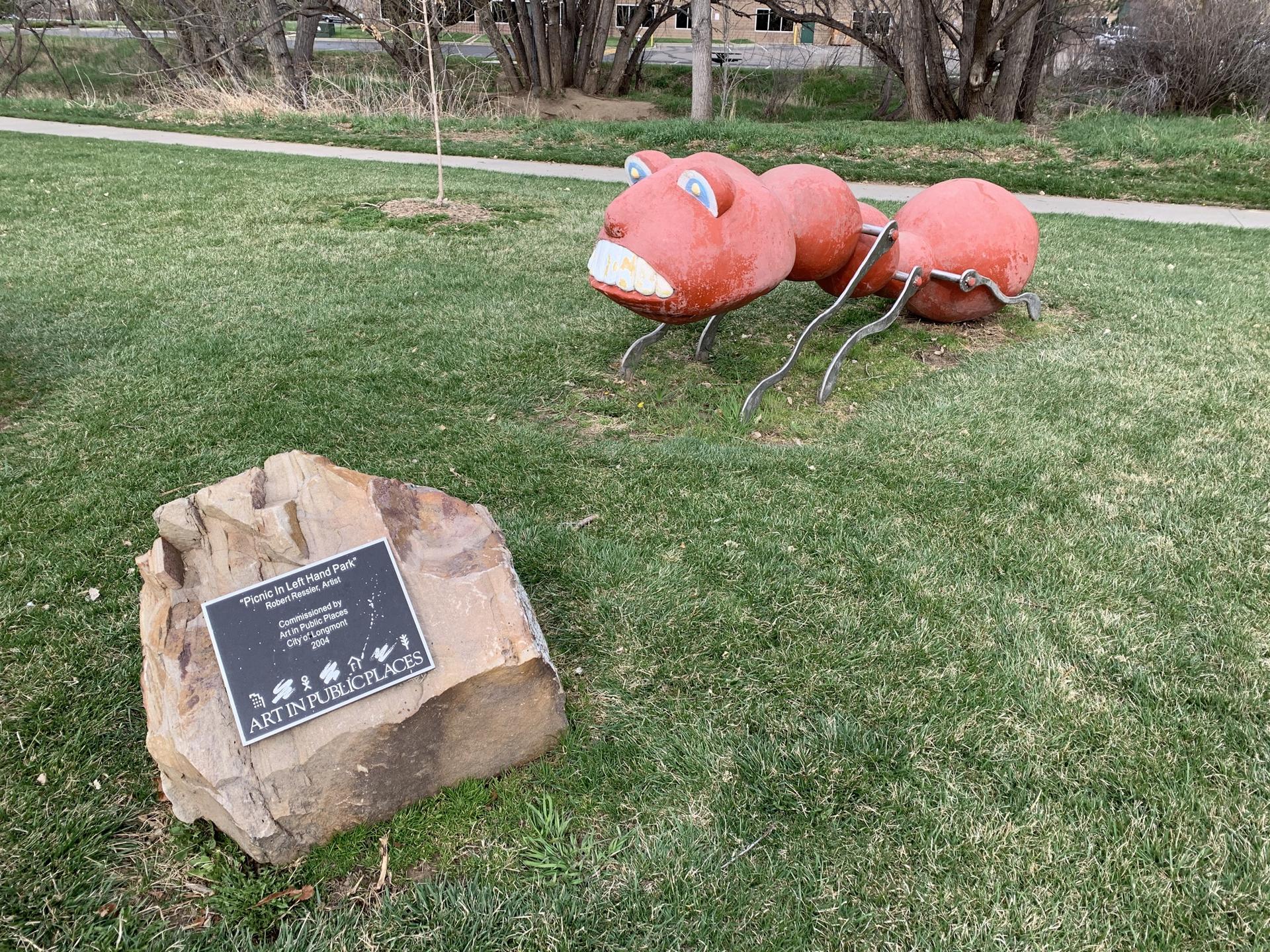 Red ant sculpture on green grass next to a rock with a plaque.
