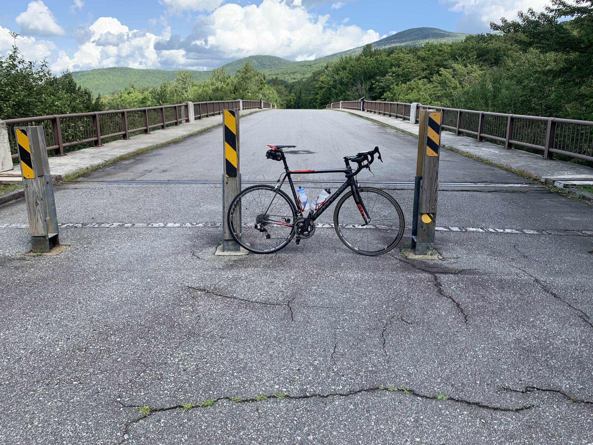 A black bicycle rests on a paved road with a bridge and mountains. - Franconia Notch Bike Path loop