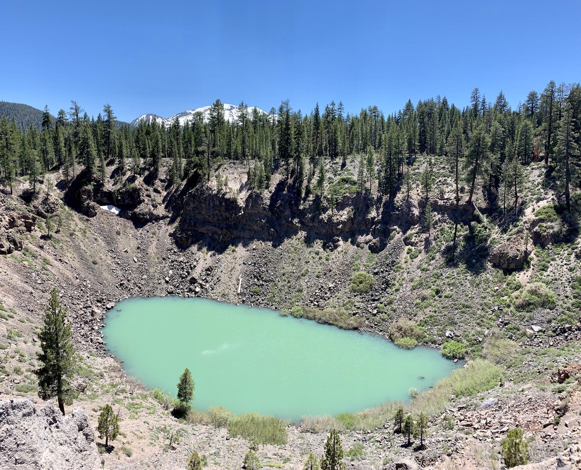Light green lake in a deep crater, surrounded by forest and mountains.