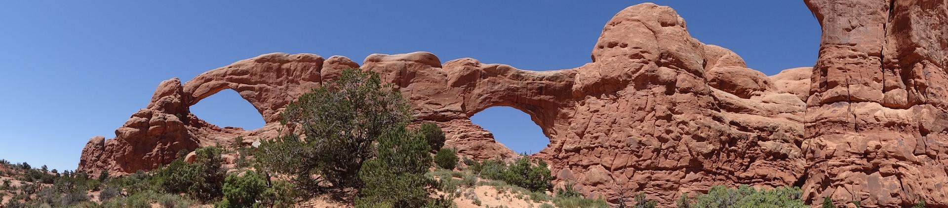 Panoramic view of two large natural arches in red rock, sunny day.