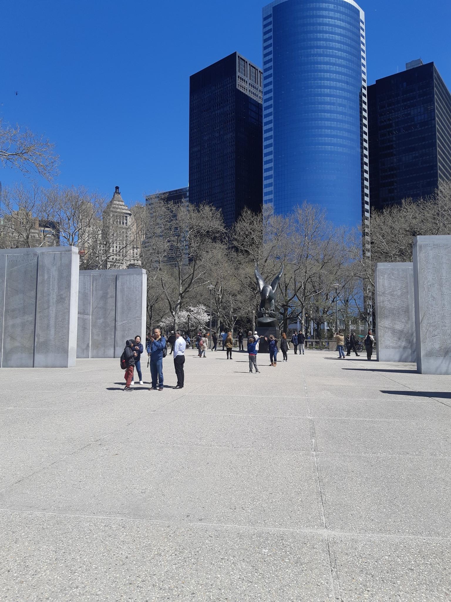 People walking in a plaza with tall buildings and trees under a clear sky.