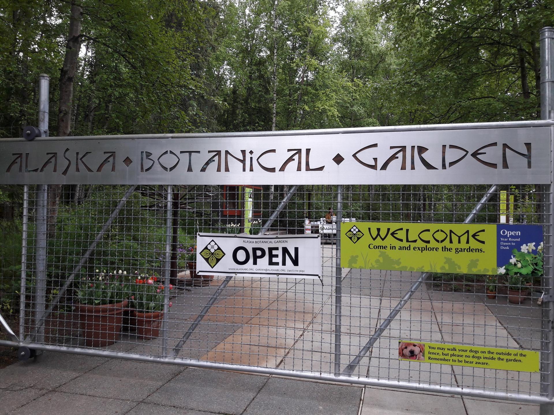 Metal gate with a large sign, "OPEN" and "WELCOME" banners, green trees.