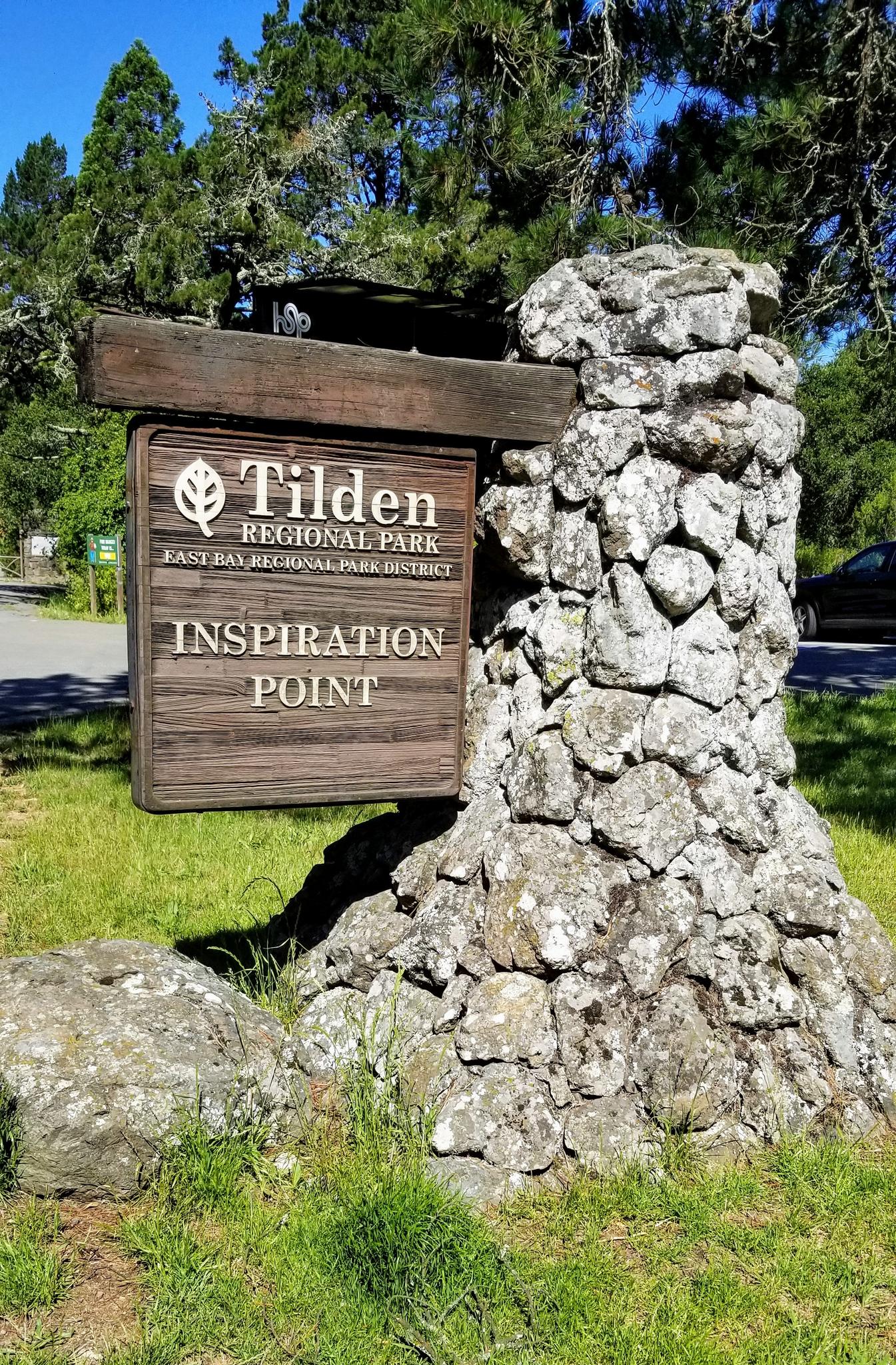Wooden sign on stone pillar in sunny, grassy park entrance.