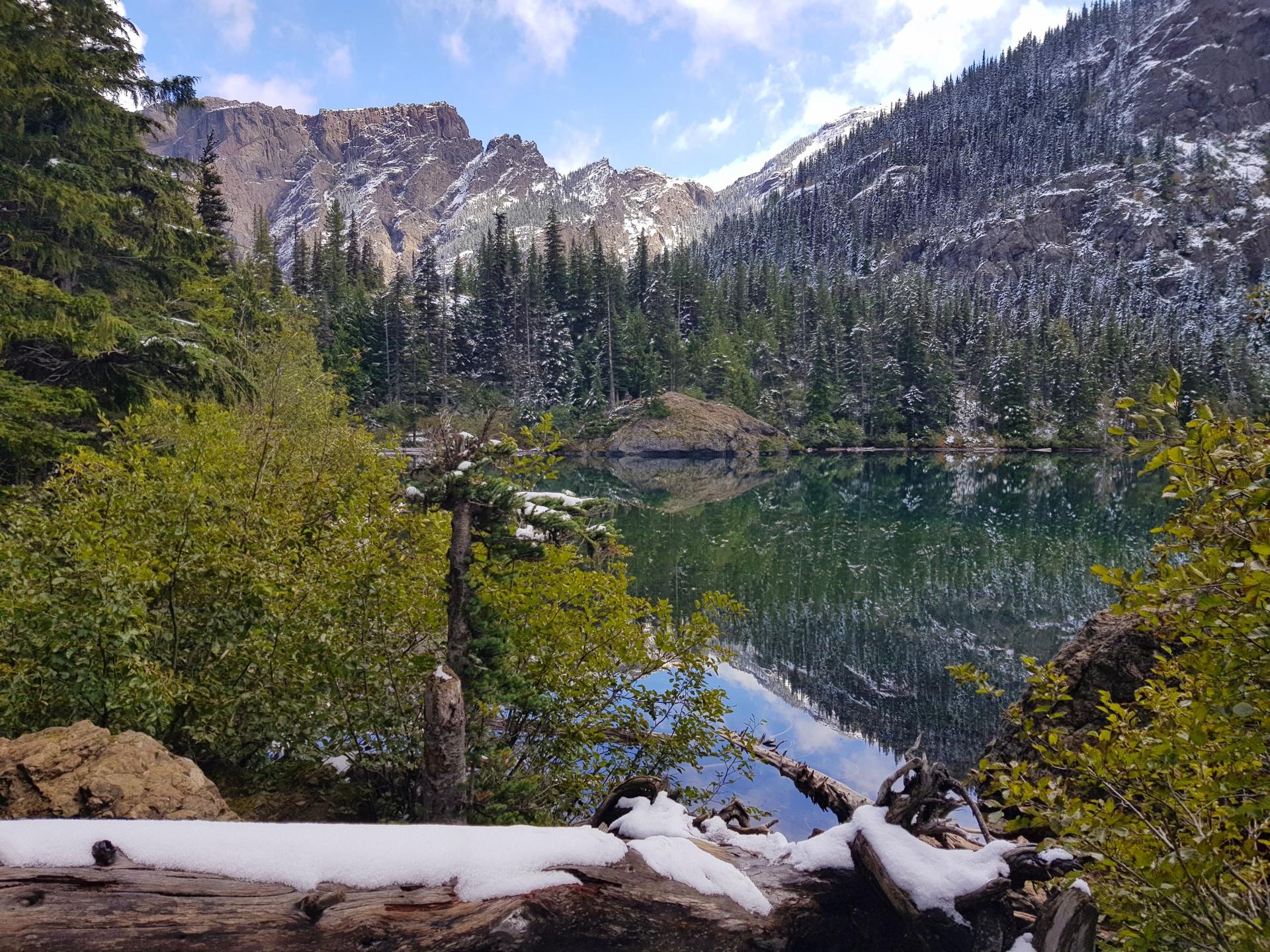 Clear lake reflecting snowy mountains and evergreen trees under a bright sky.